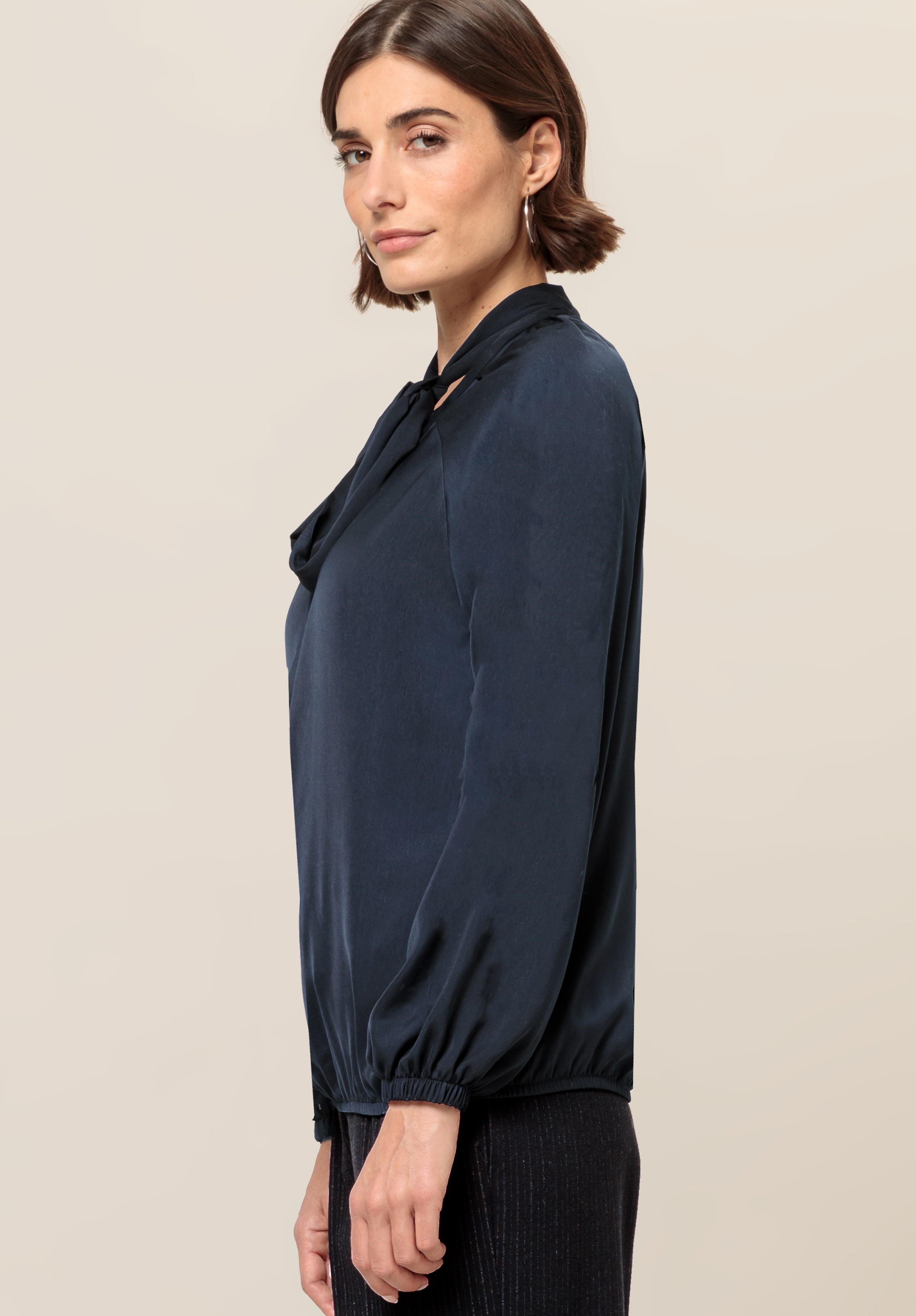 Frau mit kurzen braunen Haaren, die eine marineblaue Langarmbluse mit Schleifendetail trägt, steht seitlich und blickt vor einem neutralen Hintergrund in die Kamera.