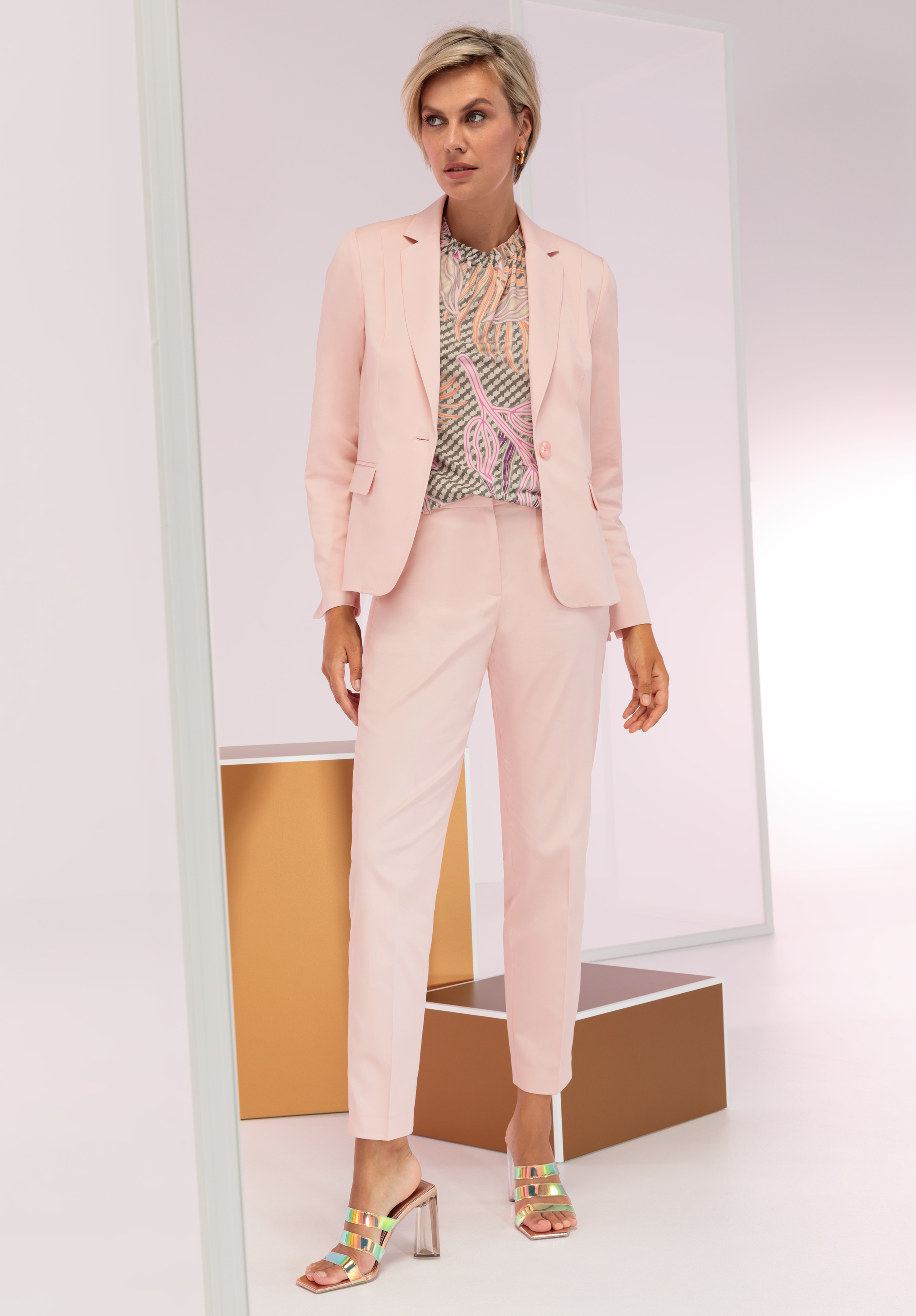 Eine Frau steht in einem minimalistischen Studio. Sie trägt einen hellrosa Hosenanzug, eine gemusterte Bluse und metallische offene Pumps. Sie hat kurze blonde Haare und eine selbstbewusste Pose, umgeben von geometrischen Requisiten.