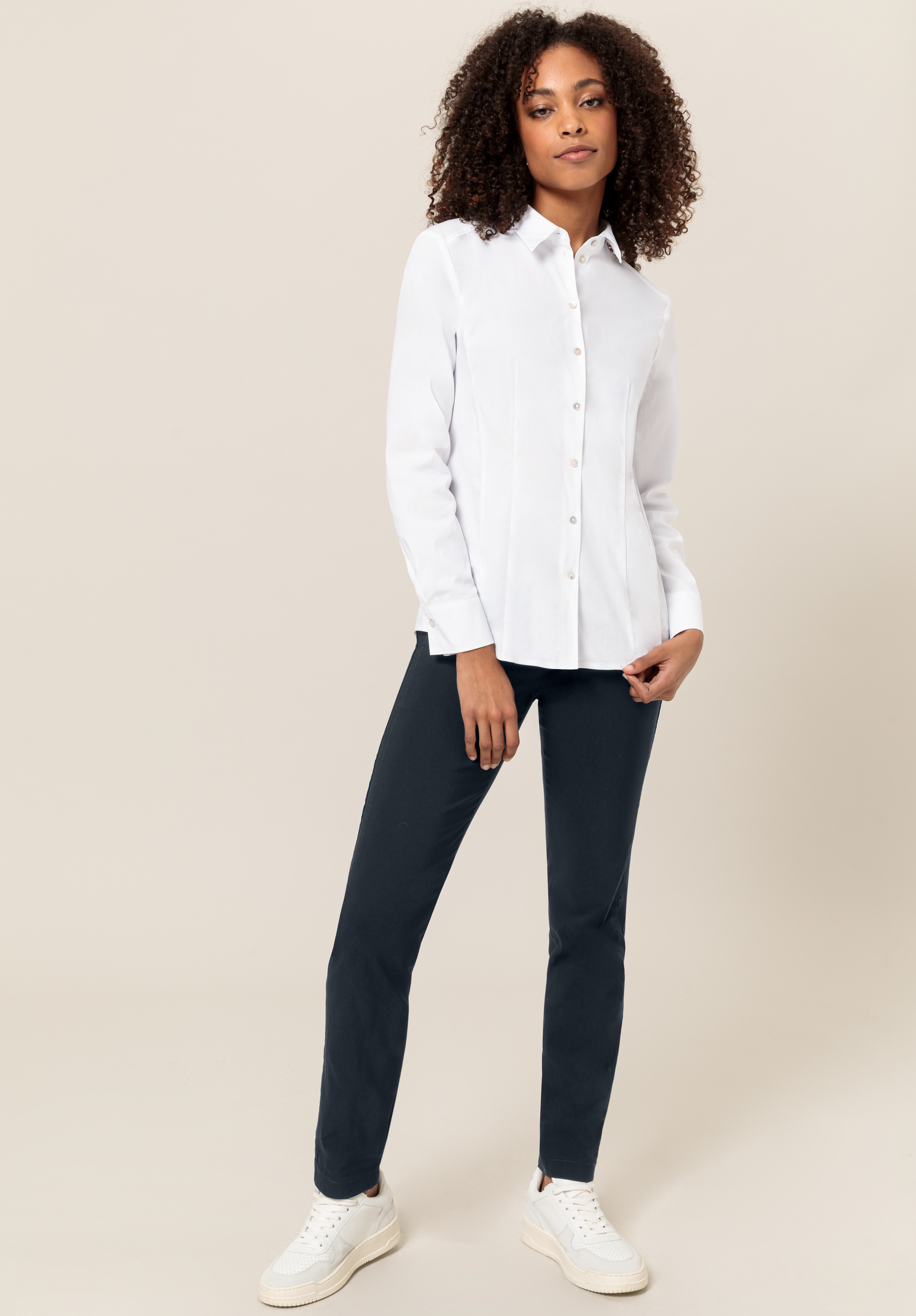 Eine Frau mit lockigem Haar steht selbstbewusst da, trägt ein weißes Button-up-Hemd, eine dunkle Hose und weiße Turnschuhe und posiert vor einem hellbeigen Hintergrund.