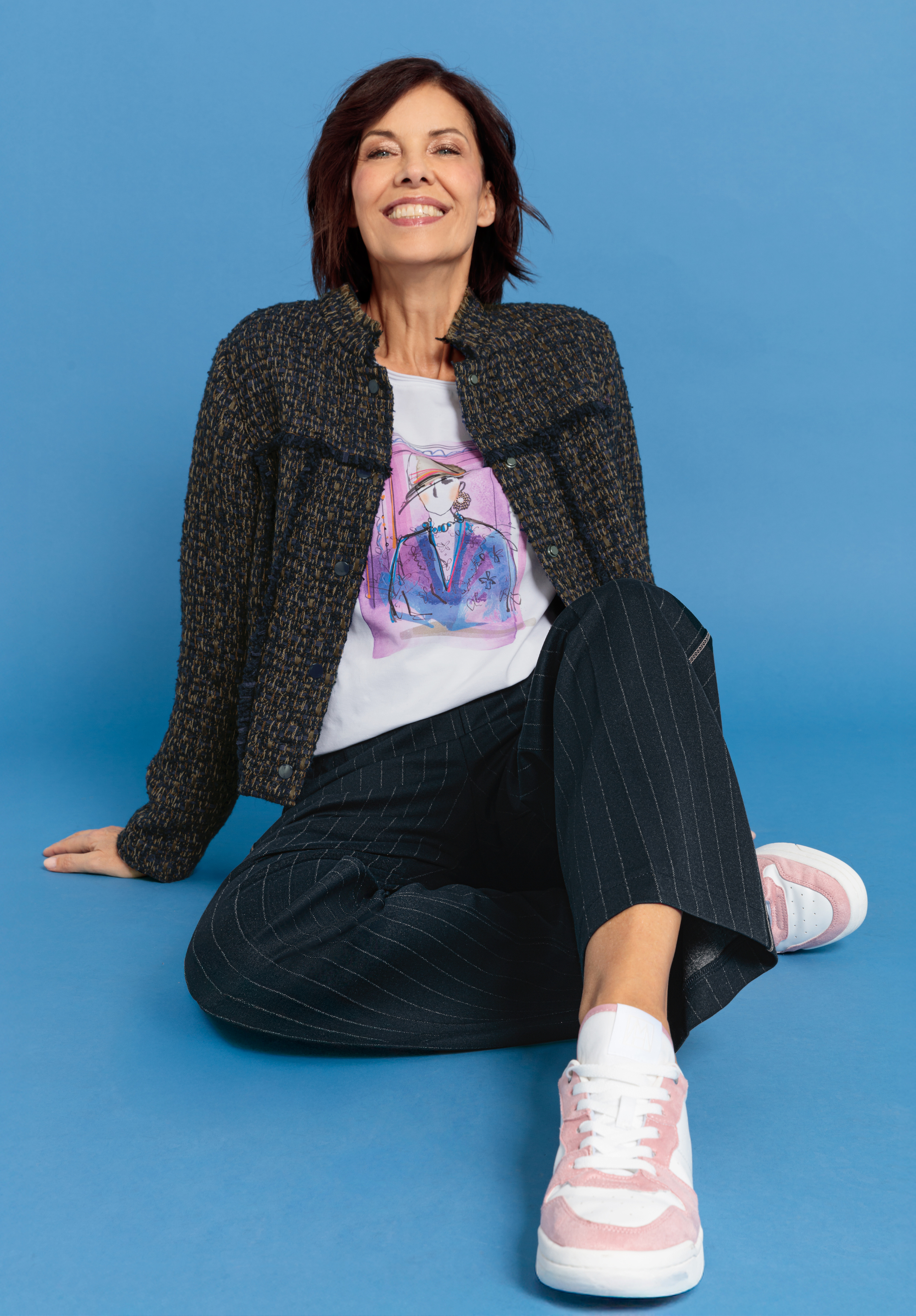 Eine lächelnde Frau mit kurzen braunen Haaren sitzt auf dem Boden vor einem blauen Hintergrund und trägt ein bedrucktes T-Shirt, eine dunkle strukturierte Jacke, eine Nadelstreifenhose und rosa-weiße Turnschuhe.