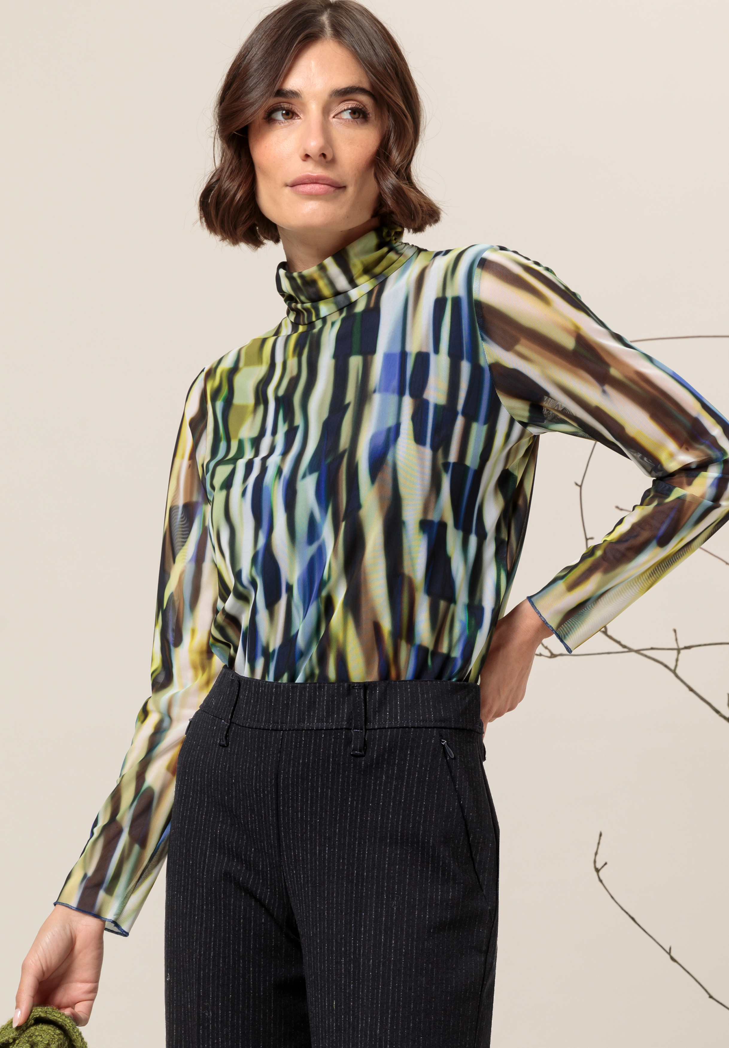 Print-Shirt GRETA Eine Frau mit kurzen braunen Haaren trägt eine bunte Rollkragenbluse mit abstraktem Muster und hoch taillierte, dunkle Nadelstreifenhosen und steht mit einer Hand auf der Hüfte vor einem neutralen Hintergrund mit sichtbaren kahlen Ästen.