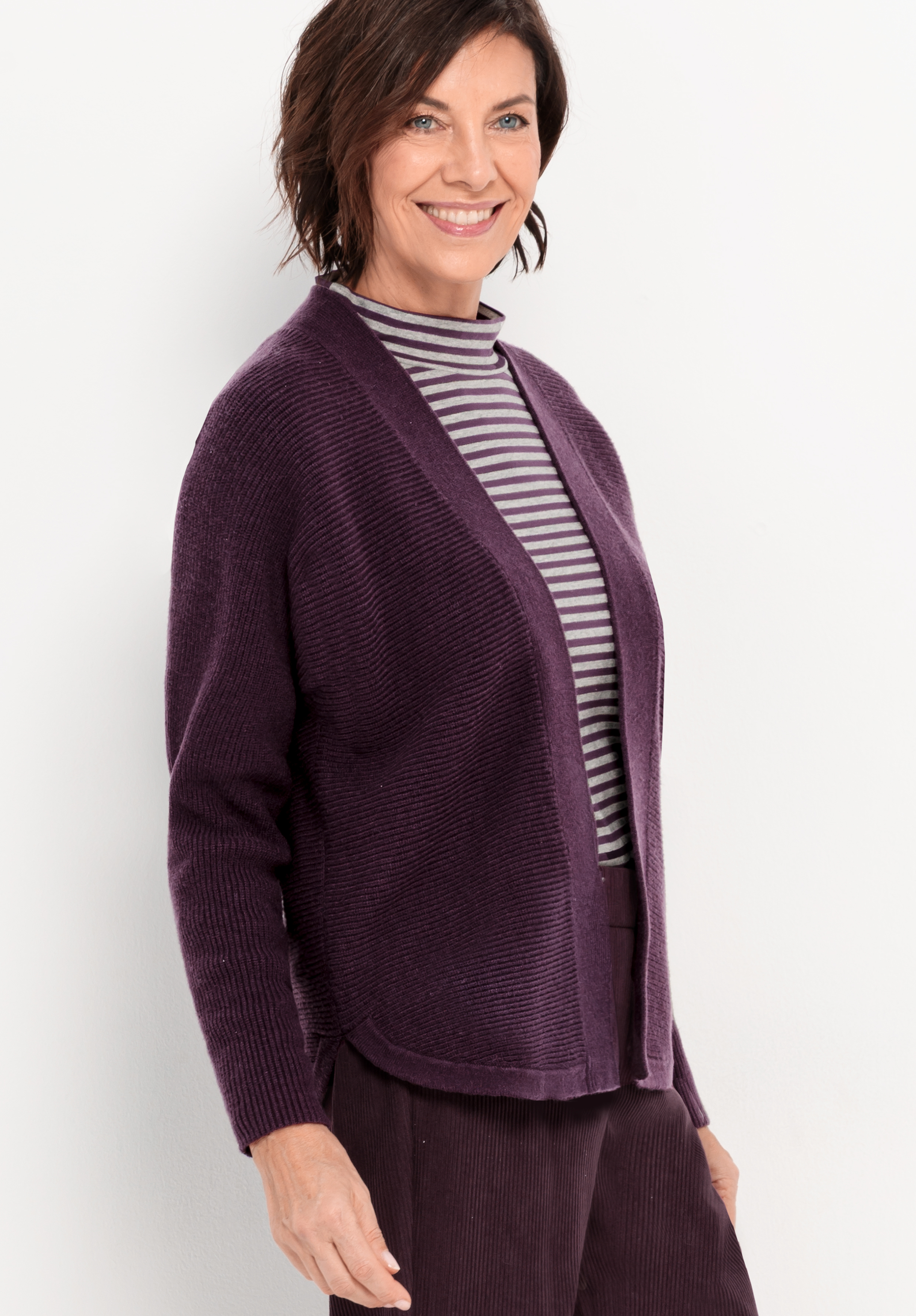 Eine Frau mit kurzen braunen Haaren lächelt, während sie eine lilafarbene Strickjacke über einem grau-weiß gestreiften Rollkragenpullover und eine dazu passende lilafarbene Hose vor einem schlichten hellen Hintergrund trägt.