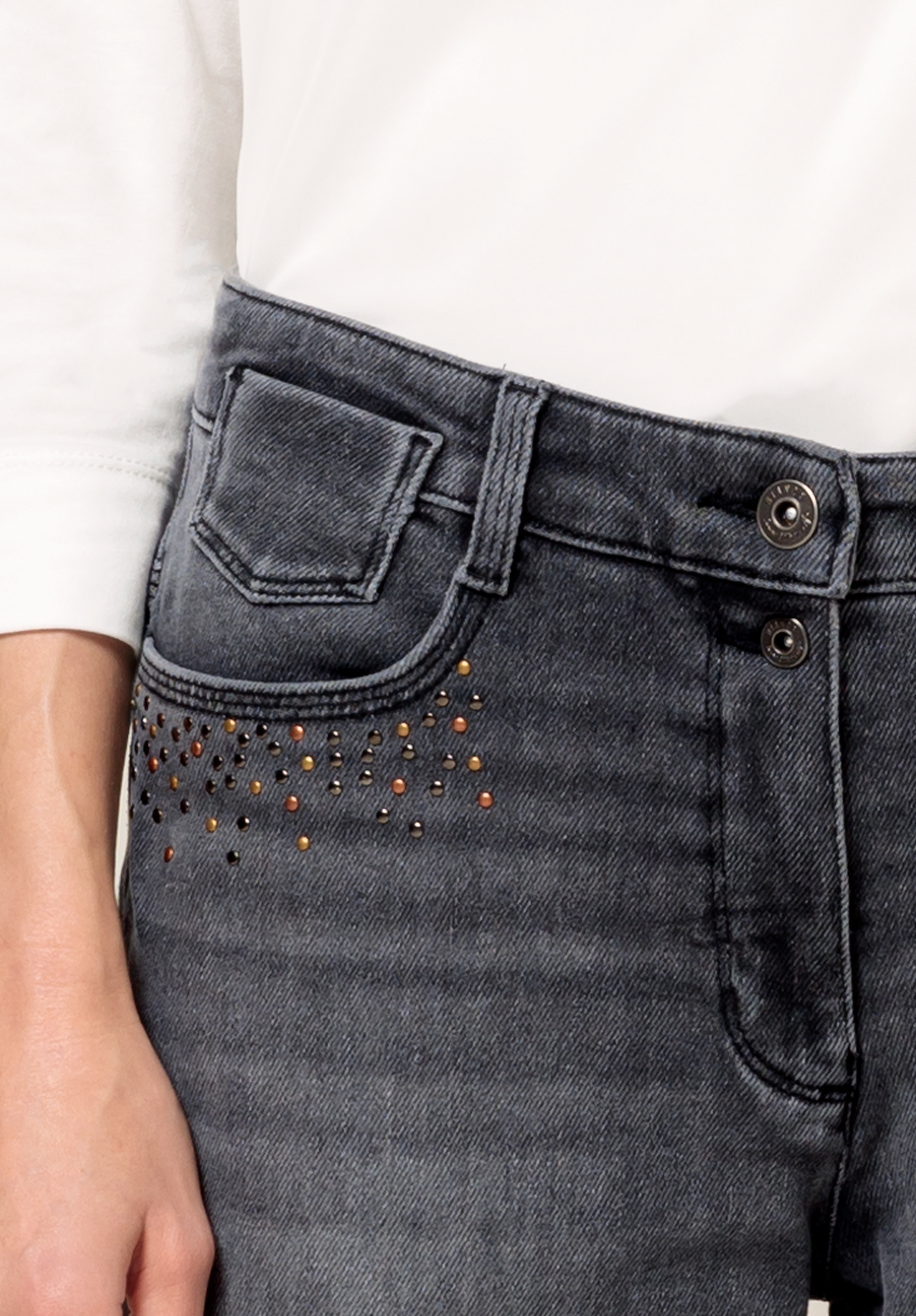 Eine Nahaufnahme einer Person, die hoch taillierte, dunkelgraue Jeans mit metallischen Nietenverzierungen in der Nähe der Tasche und ein weißes Langarmshirt trägt. Nur ein Teil des Arms und der Hand ist sichtbar.