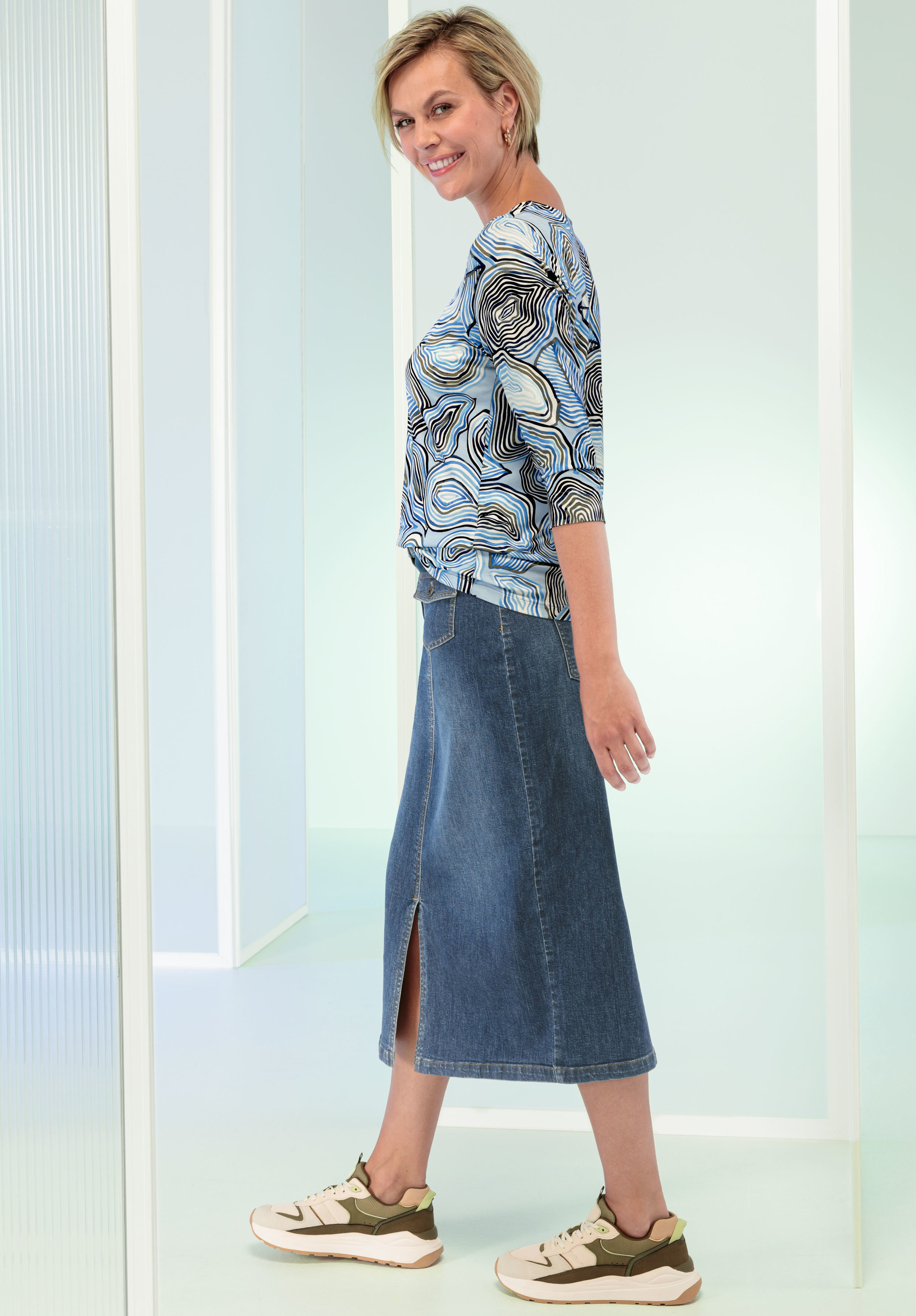 Eine Frau mit kurzen blonden Haaren lächelt beim Gehen. Sie trägt ein gemustertes blaues Oberteil, einen knielangen Jeansrock mit Schlitz vorne und beige Sneakers mit grünen Akzenten in einem modernen, lichtdurchfluteten Raum.