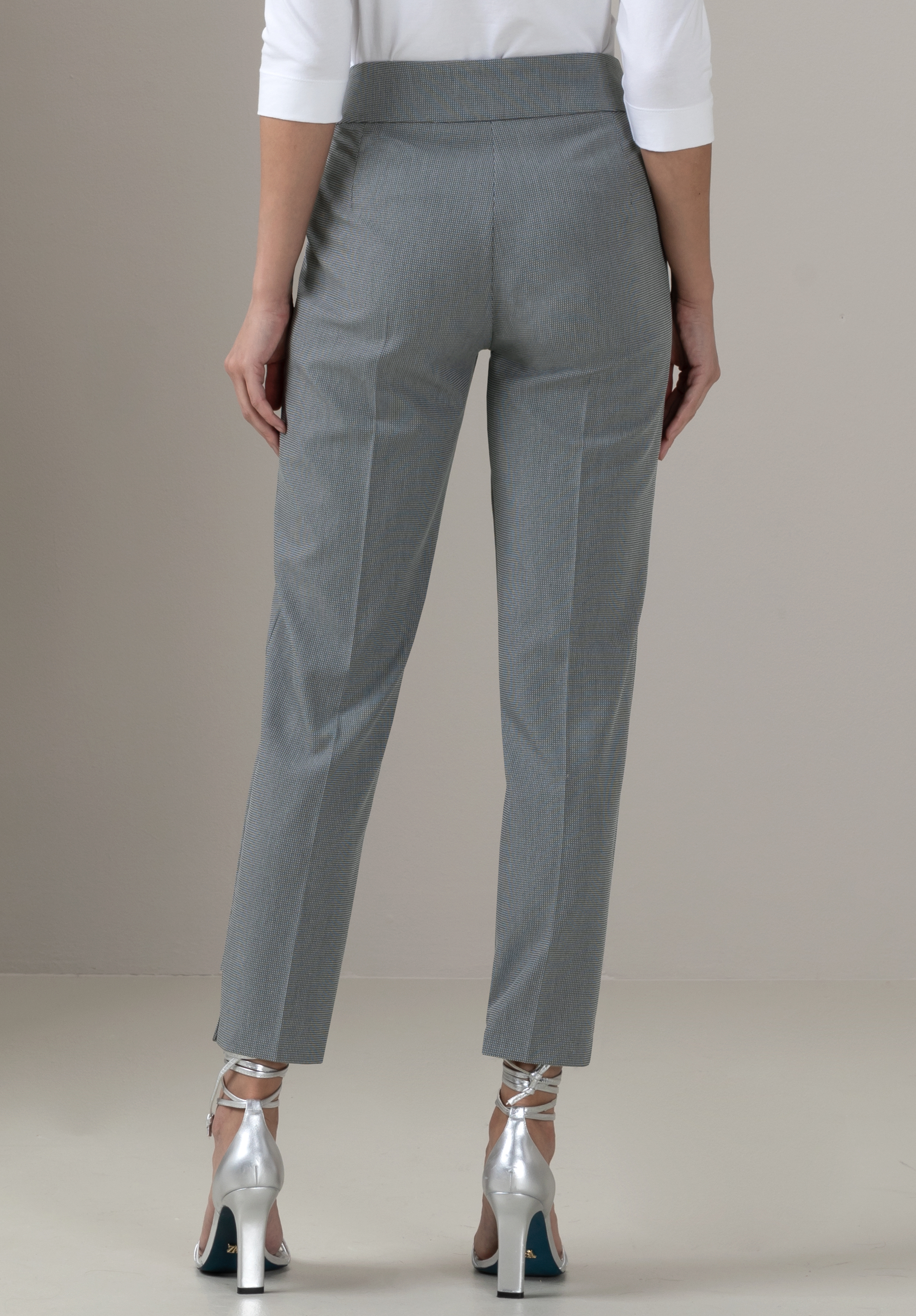 Eine Person, die eine graue Anzughose, ein weißes Oberteil und silberne hochhackige Schuhe trägt, steht mit dem Rücken zur Kamera vor einem schlichten hellen Hintergrund.