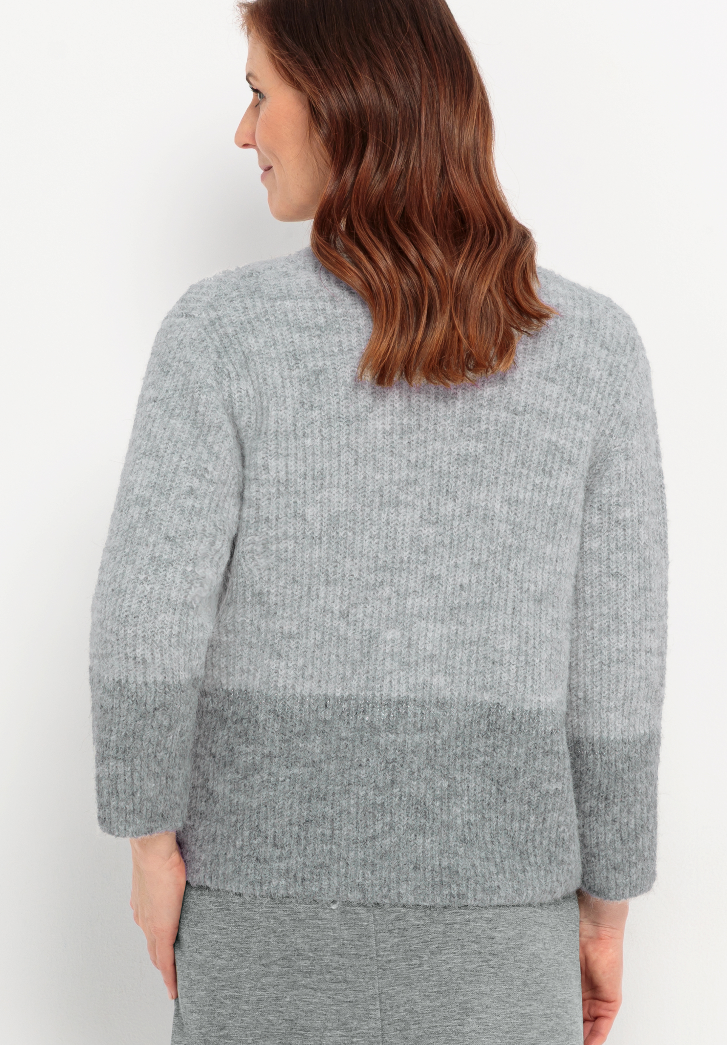 Eine Frau mit schulterlangen braunen Haaren steht mit dem Gesicht nach hinten und trägt einen hellgrauen, langärmeligen, strukturierten Pullover mit einem dunkleren grauen Abschnitt am unteren Ende und einen dazu passenden grauen Rock.