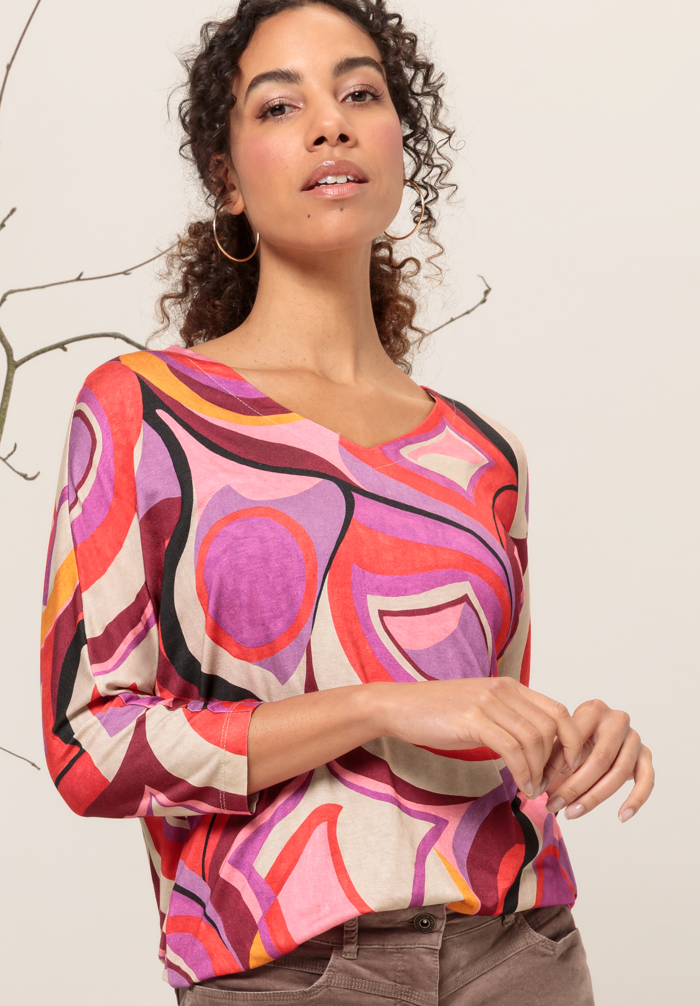 Eine Frau mit lockigem Haar trägt große Creolen und ein farbenfrohes Langarmshirt mit abstraktem Muster in den Farben Rosa, Rot, Lila und Beige und steht vor einem neutralen Hintergrund mit einem kahlen Ast.
