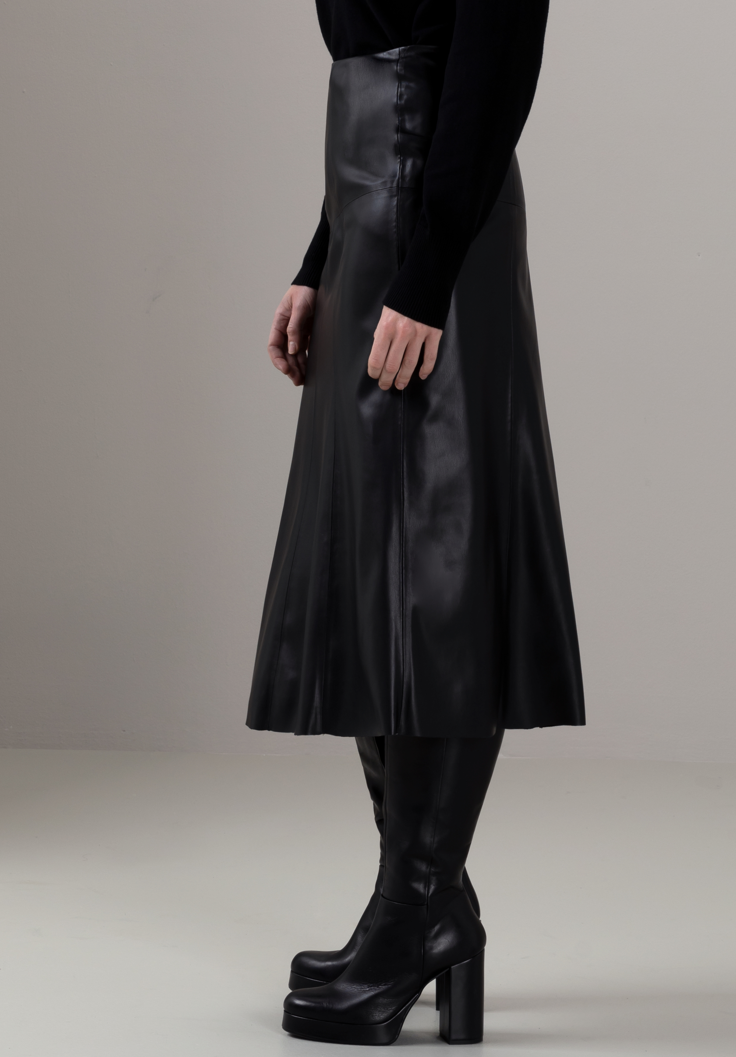 Eine Person, die ein schwarzes Langarmoberteil, einen schwarzen Midirock aus Kunstleder und schwarze kniehohe Plateaustiefel trägt, steht vor einem schlichten hellen Hintergrund.
