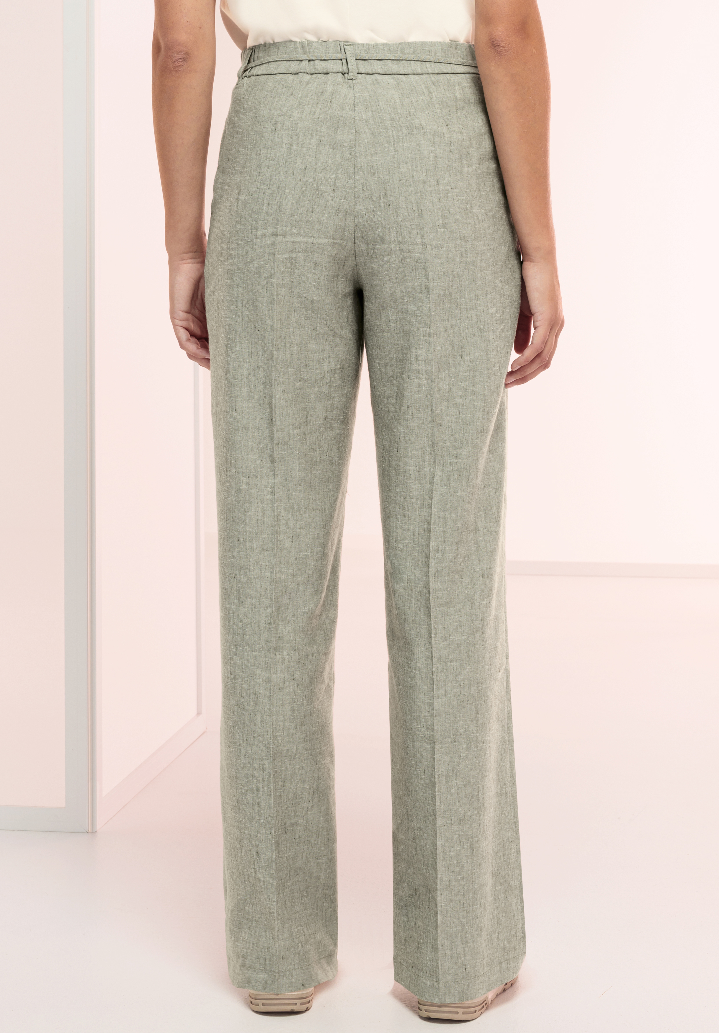 Eine Person steht im Innenbereich und ist von der Hüfte abwärts abgebildet. Sie trägt eine hoch taillierte, hellgraue Hose und beige Schuhe. Der Hintergrund ist minimalistisch und sanft beleuchtet.