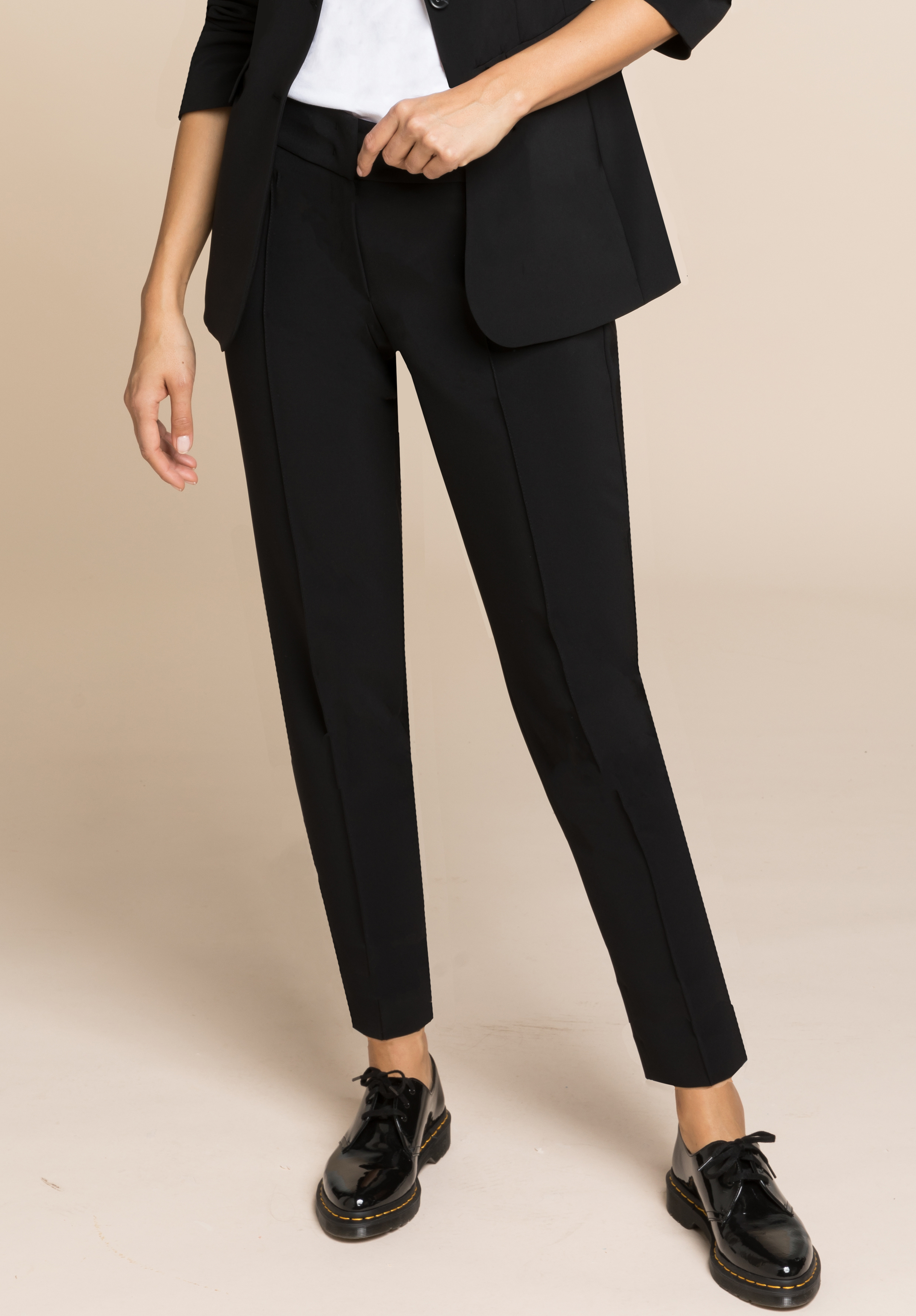 Eine Person, die eine schwarze, maßgeschneiderte Hose, einen schwarzen Blazer und schwarze, glänzende Schnürschuhe trägt, steht vor einem beigen Hintergrund. Zu sehen ist nur die untere Körperhälfte.