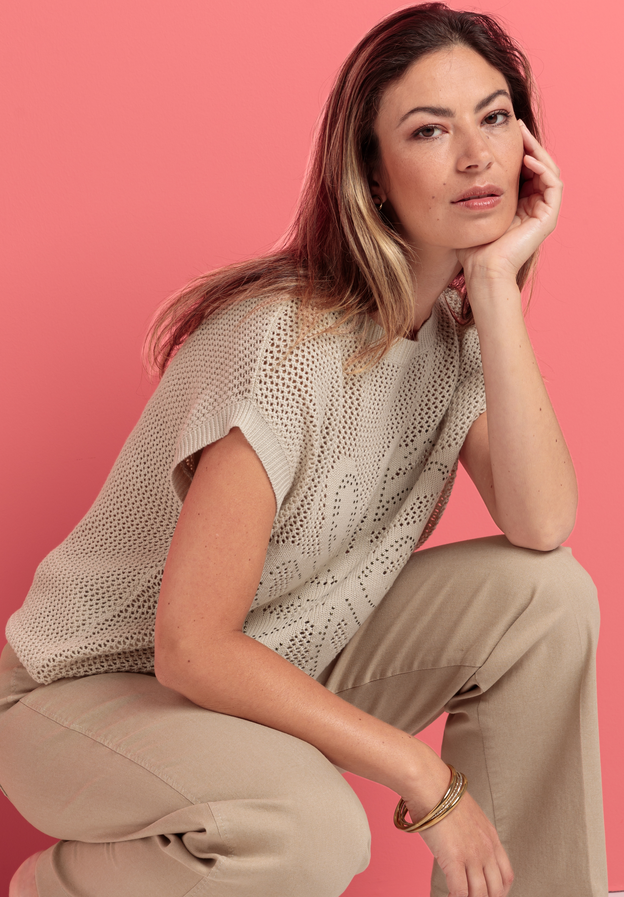 Eine Frau mit hellbraunem Haar sitzt vor einem rosa Hintergrund. Sie trägt ein beiges Strickoberteil, hellbeige Hosen und goldene Armreifen. Sie stützt ihr Kinn auf ihre Hand und blickt mit entspanntem Gesichtsausdruck in die Kamera.