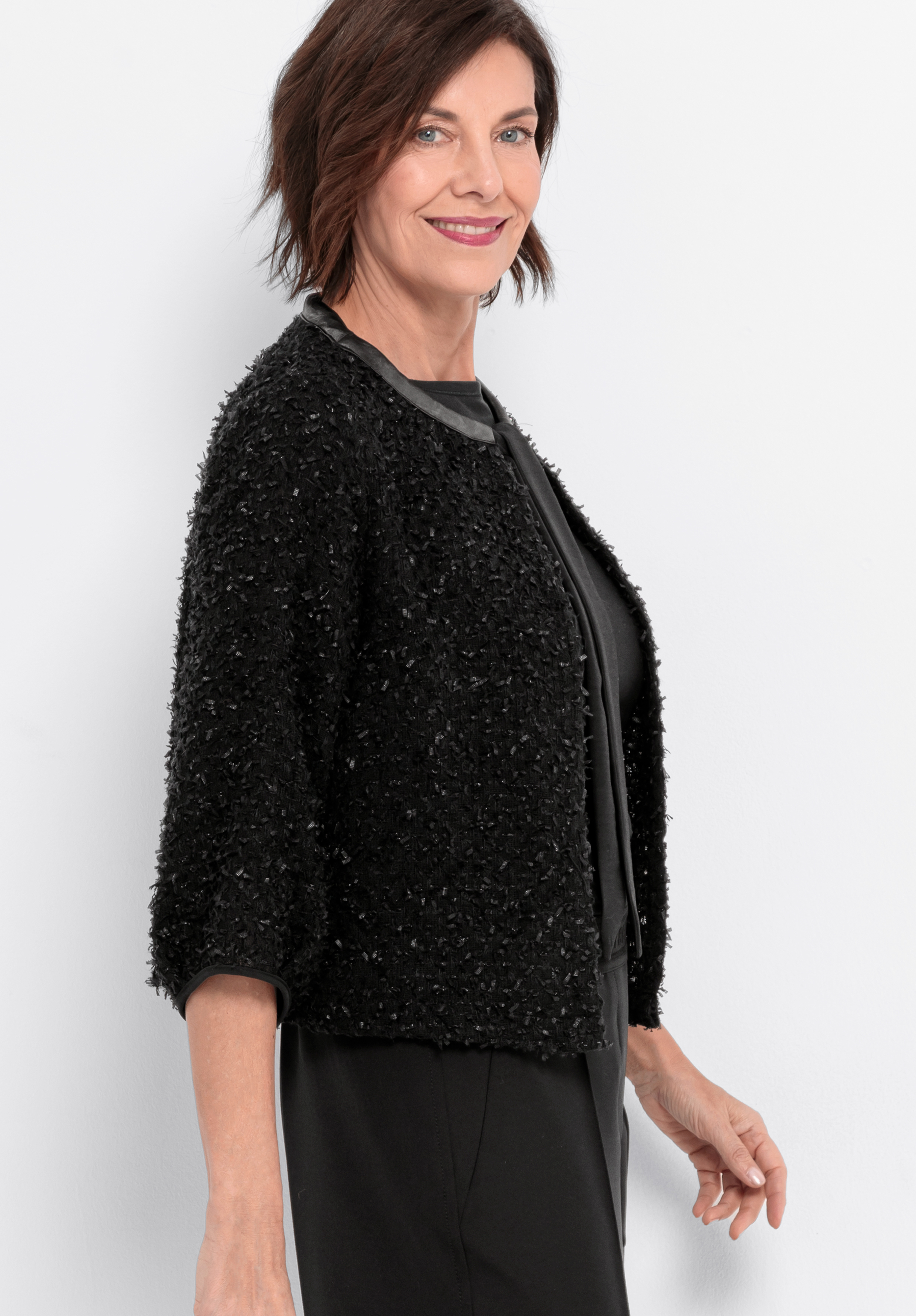 Eine Frau mit kurzen braunen Haaren, die eine strukturierte schwarze Jacke über einem schwarzen Kleid trägt, steht vor einem schlichten hellen Hintergrund, lächelt und blickt leicht zur Seite.