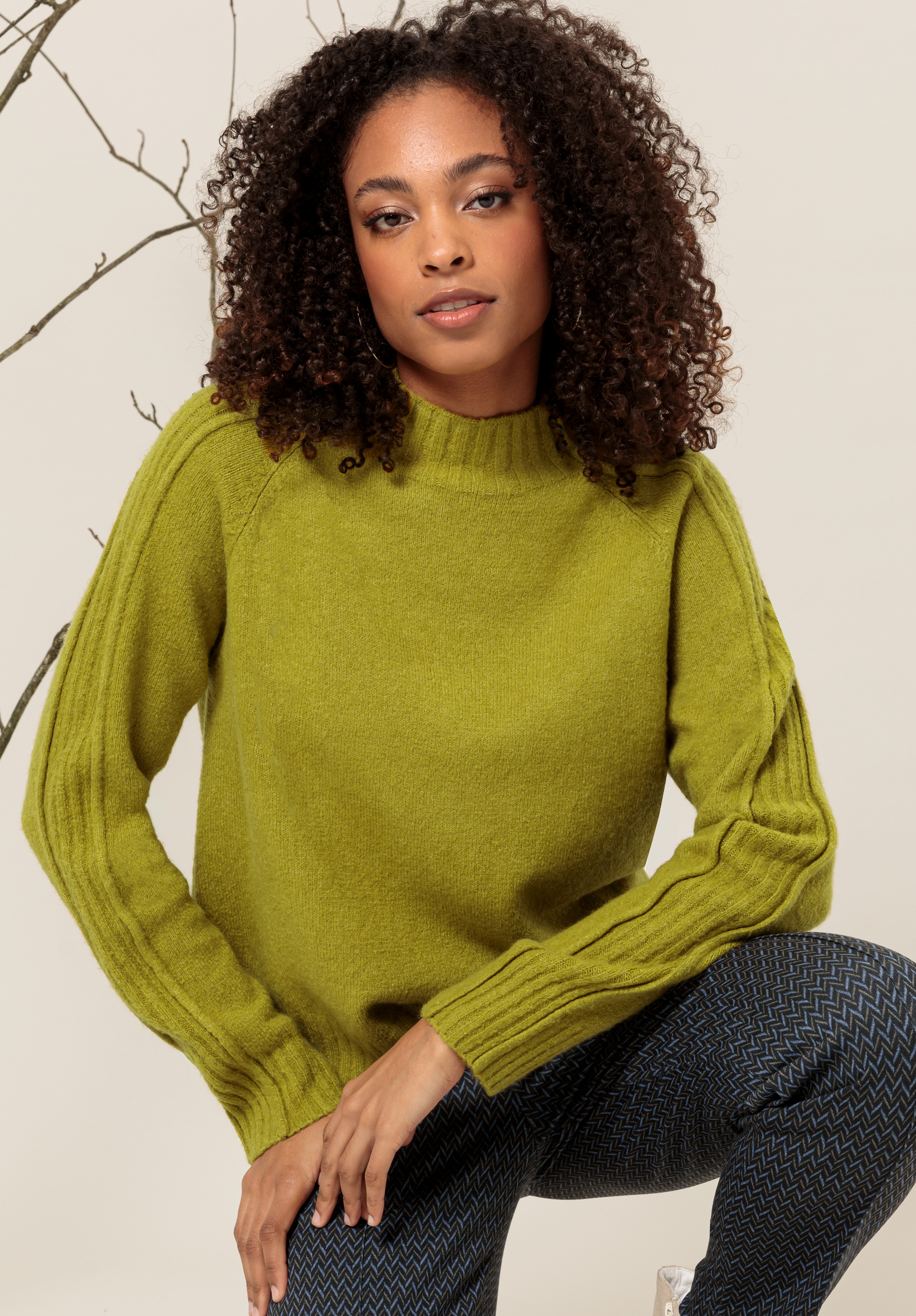 Eine Frau mit lockigem Haar, einem olivgrünen Pullover und gemusterten blauen Hosen sitzt vor einem neutralen Hintergrund, auf der linken Seite ist ein kahler Ast zu sehen.