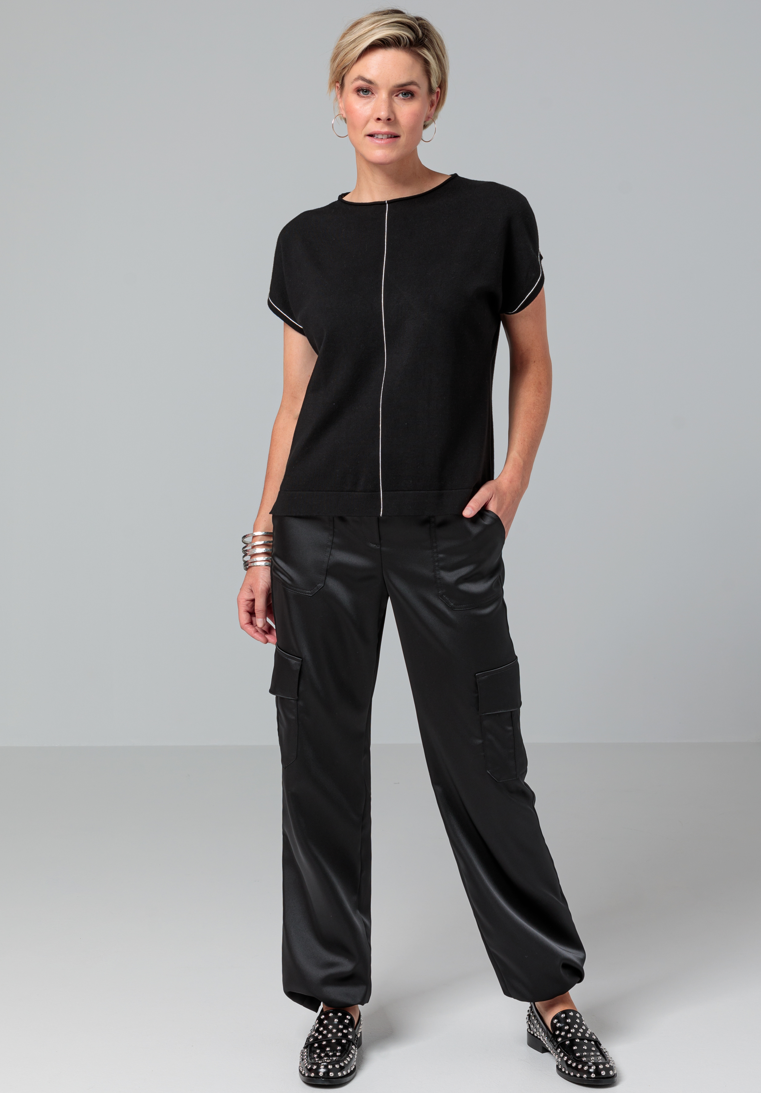 Eine Frau steht vor einem schlichten Hintergrund. Sie trägt ein schwarzes Kurzarmoberteil, schwarze Cargohosen, schwarze Schuhe mit Nieten und silberne Armbänder. Sie hat kurze, blonde Haare und eine Hand in der Tasche.