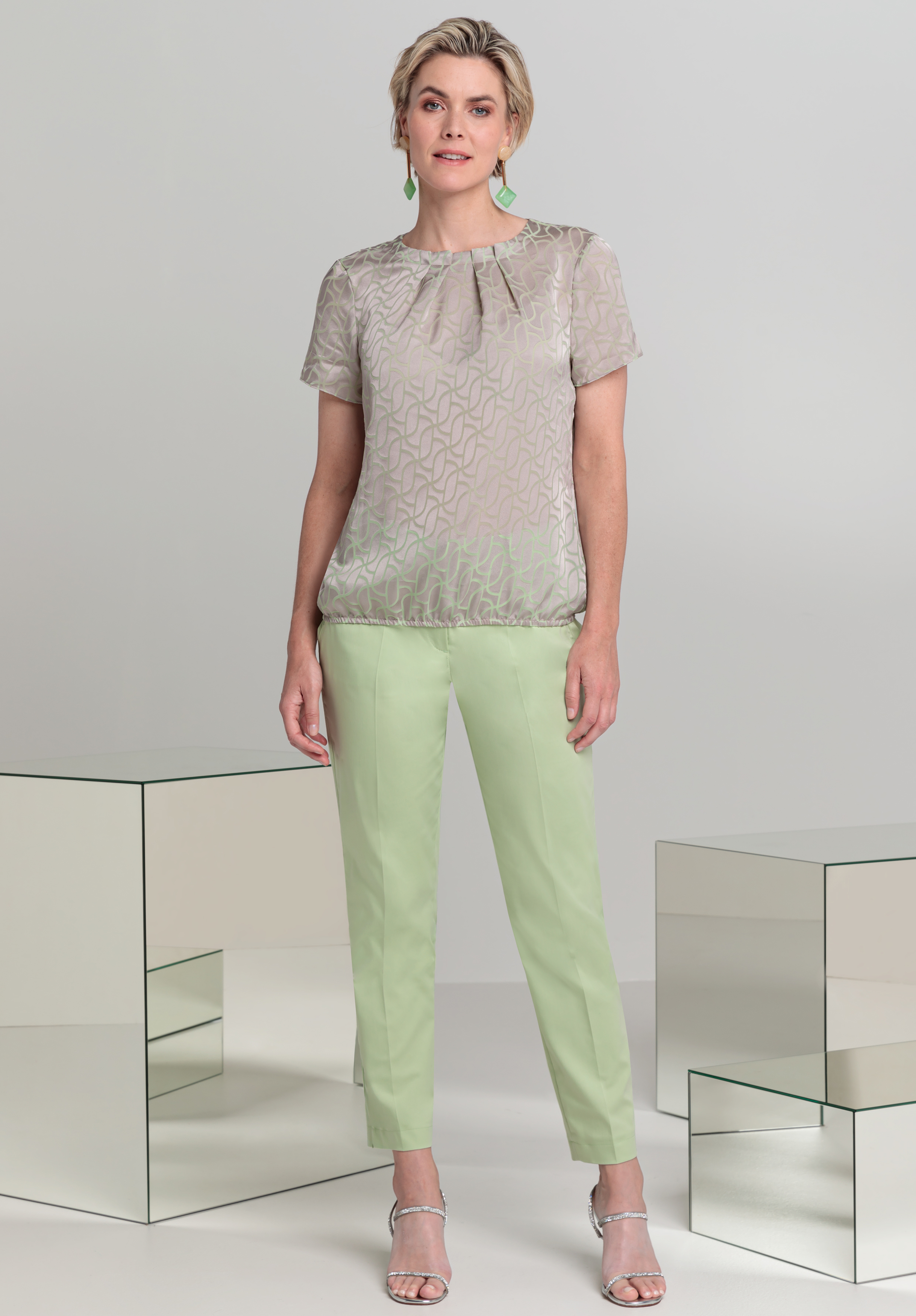 Eine Frau steht in einem Studio. Sie trägt eine kurzärmelige, gemusterte beige Bluse, hellgrüne Hosen, silberne Pumps und grüne Ohrringe. Sie ist von zwei verspiegelten kubischen Tischen umgeben.