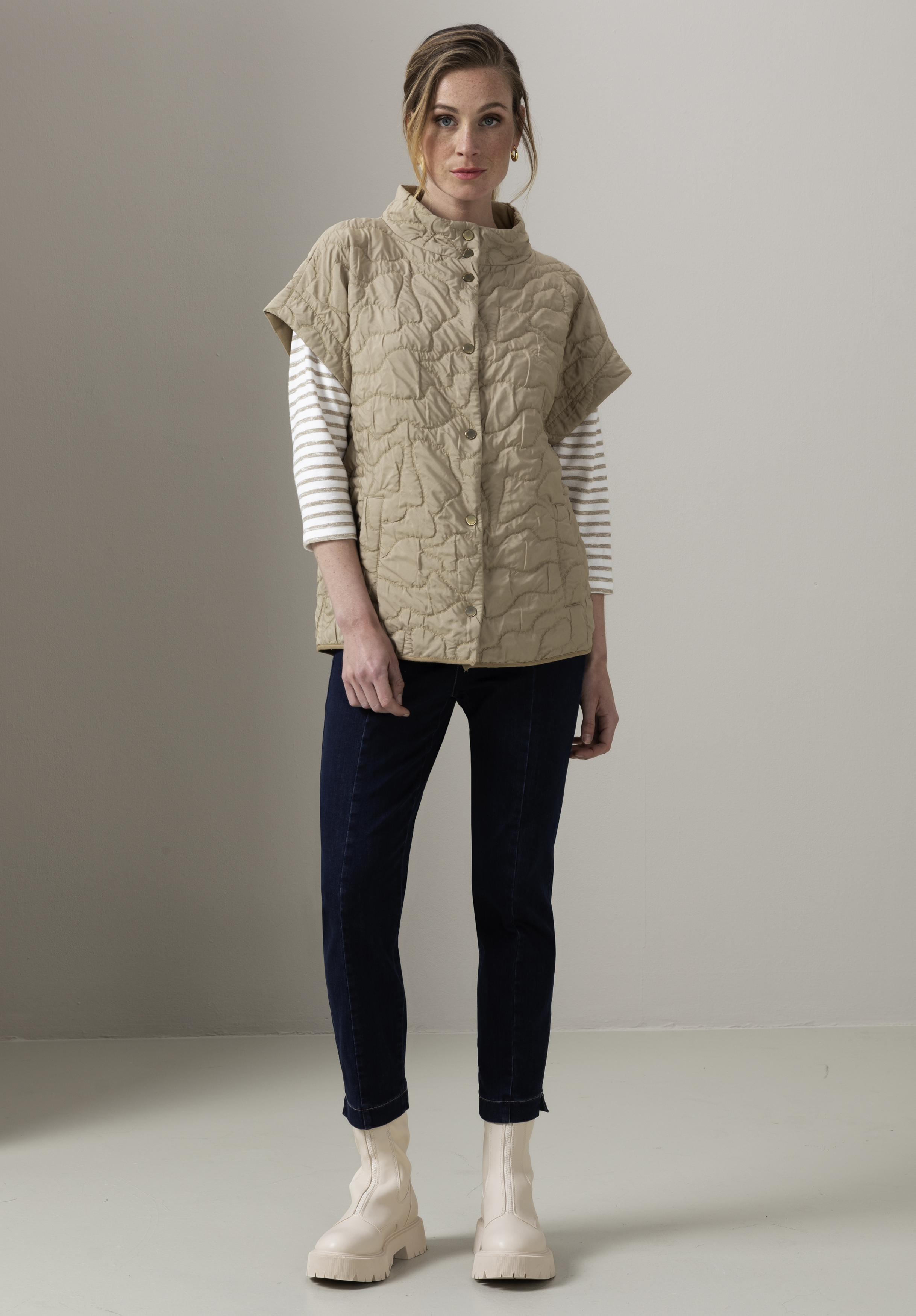 Eine Frau steht drinnen. Sie trägt eine beige gesteppte Kurzarmjacke über einem weiß-grau gestreiften Langarmshirt, dunkelblaue Jeans und hellbeige klobige Stiefel. Sie hat helles Haar zurückgekämmt und blickt in die Kamera.