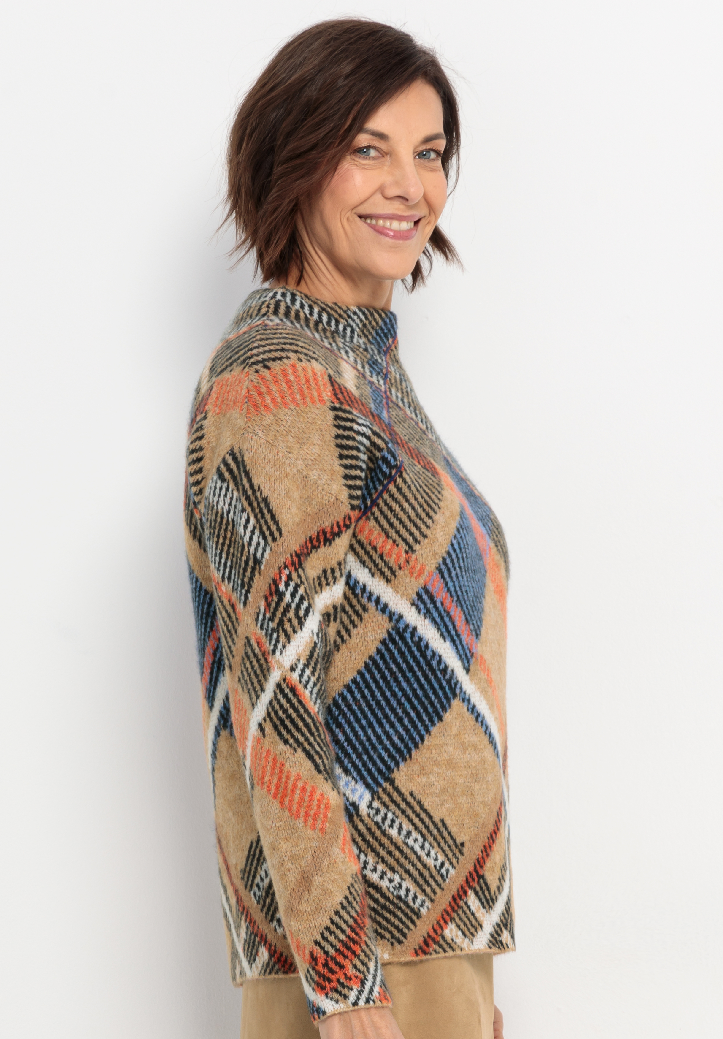 Eine Frau mit kurzen braunen Haaren lächelt, während sie seitwärts steht. Sie trägt einen beige, orange, blau und schwarz karierten Pullover und eine hellbraune Hose vor einem schlichten weißen Hintergrund.