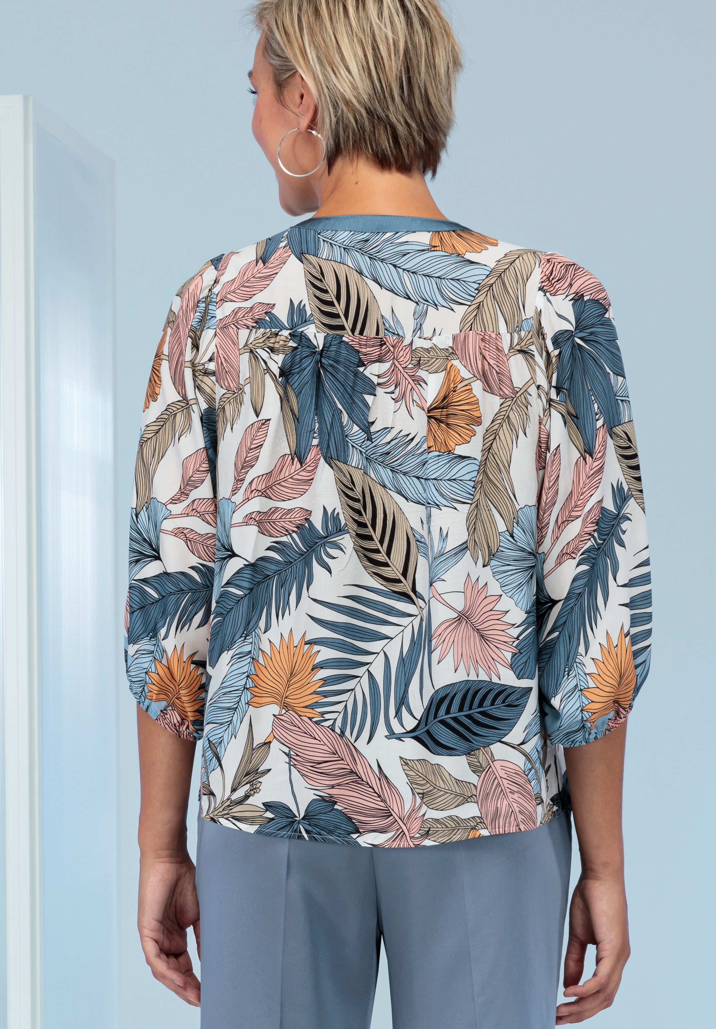 Eine Person mit kurzen, hellen Haaren ist von hinten zu sehen. Sie trägt eine Bluse mit einem farbenfrohen, tropischen Blattmuster in Blau-, Beige-, Rosa- und Schwarztönen, dazu eine hellblaue Hose. Der Hintergrund ist hellblau.