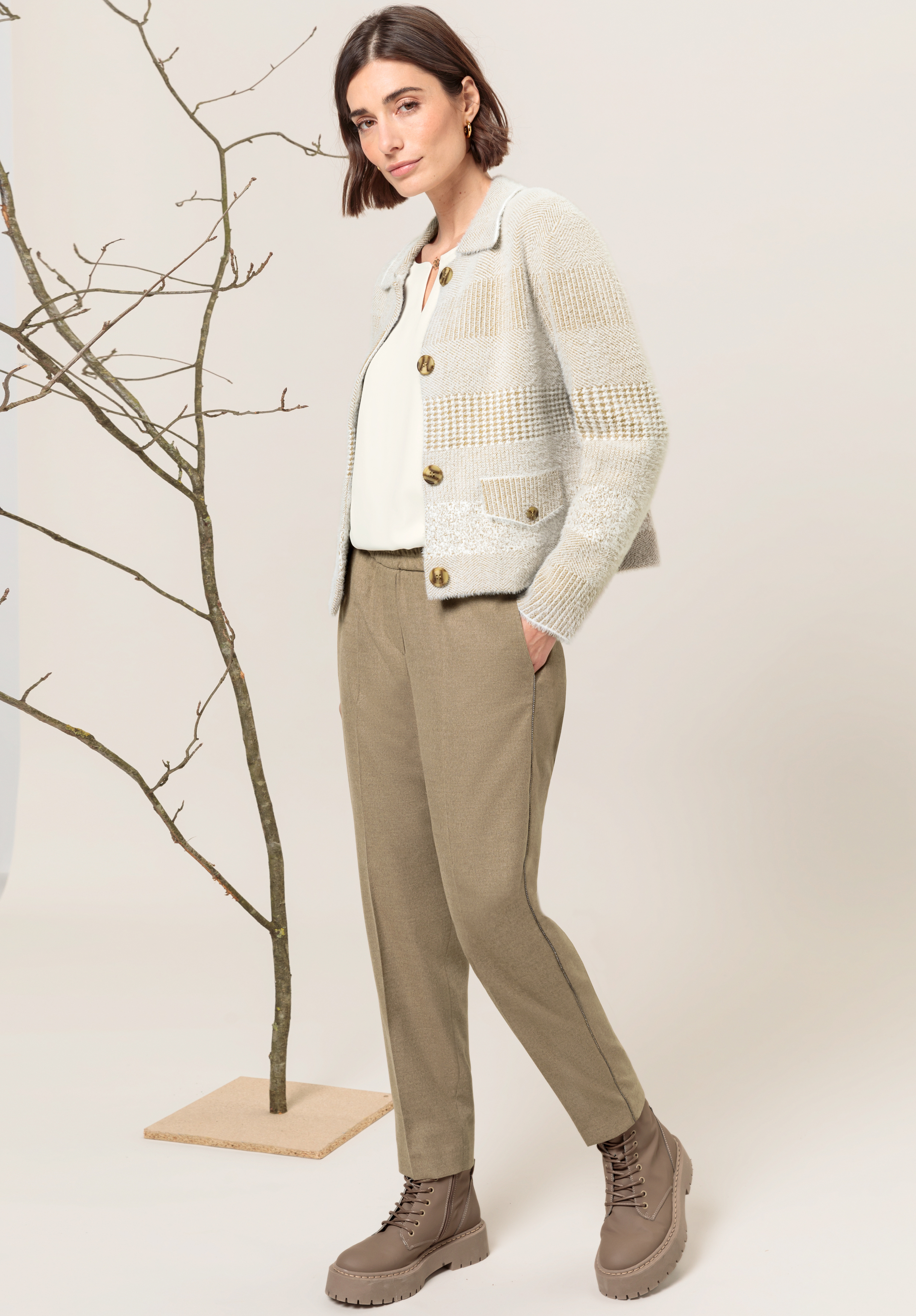 Eine Frau steht neben einem kahlen Ast. Sie trägt eine creme- und beige gestreifte Strickjacke, eine weiße Bluse, hellbraune Hosen und taupefarbene Schnürstiefel. Ihr Gesichtsausdruck ist neutral, und das Licht ist sanft und natürlich.