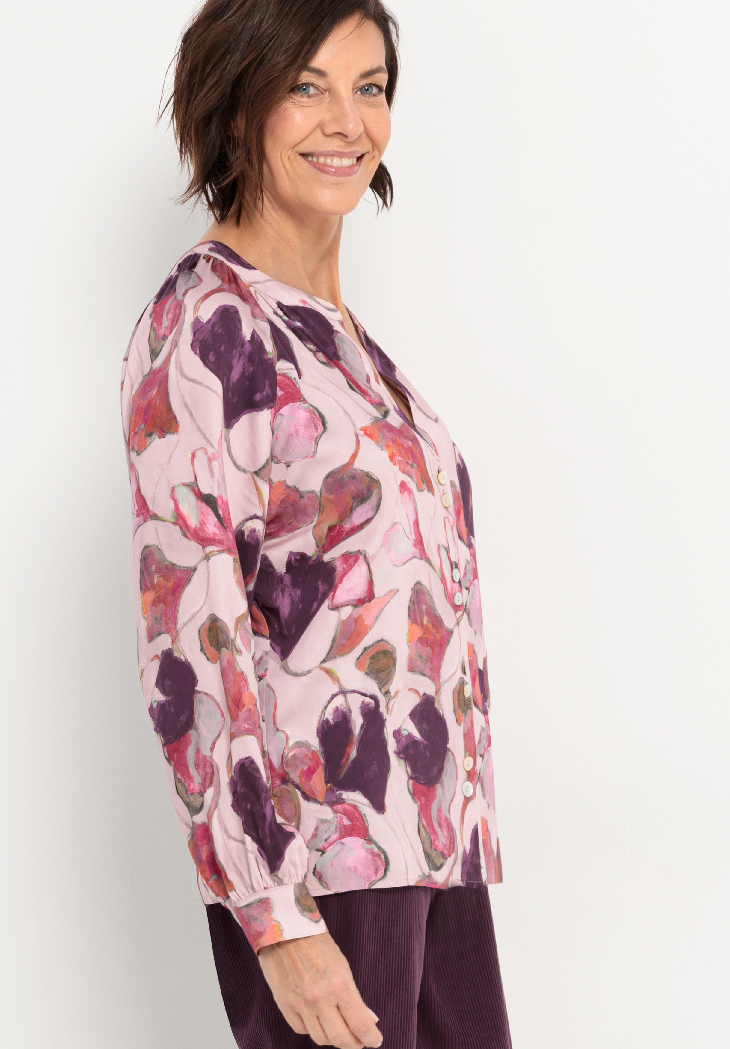 Eine Frau mit kurzen braunen Haaren lächelt, während sie eine hellrosa Bluse mit einem violetten und magentafarbenen Blumenmuster und eine passende violette Hose vor einem schlichten weißen Hintergrund trägt.