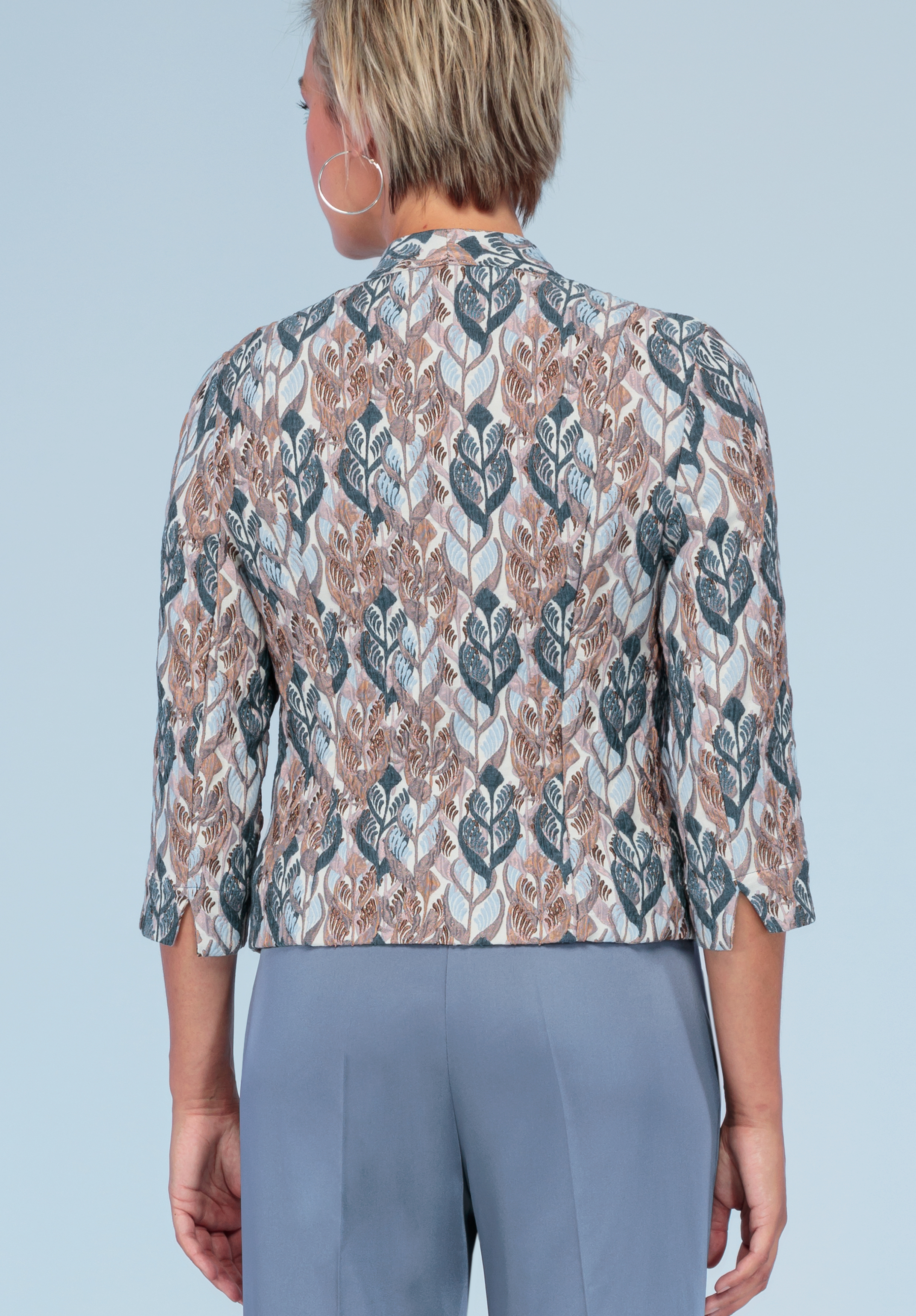 Eine Person trägt eine gemusterte Jacke mit Dreiviertelärmeln und Blättermuster in Beige-, Weiß- und Blautönen, kombiniert mit einer hellblauen Hose, von hinten auf hellblauem Hintergrund abgebildet.