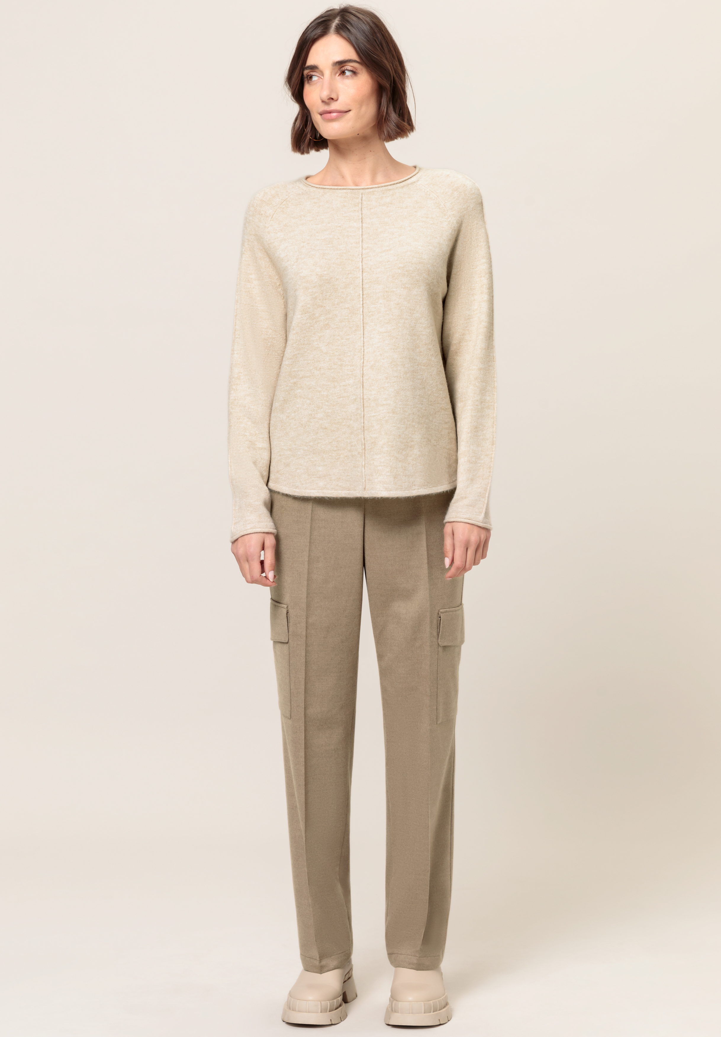 Eine Frau steht mit Blick nach vorne vor einem schlichten hellen Hintergrund und trägt einen beigen Langarmpullover, hellbraune Cargohosen und beige Schuhe.