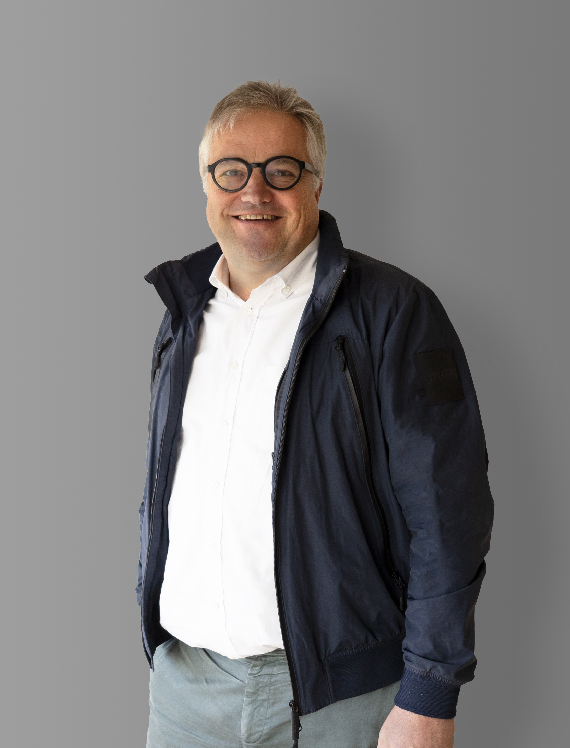 Ein lächelnder Mann mit grauem Haar und Brille trägt ein weißes Hemd, eine marineblaue Jacke und graue Hosen und steht vor einem schlichten grauen Hintergrund.