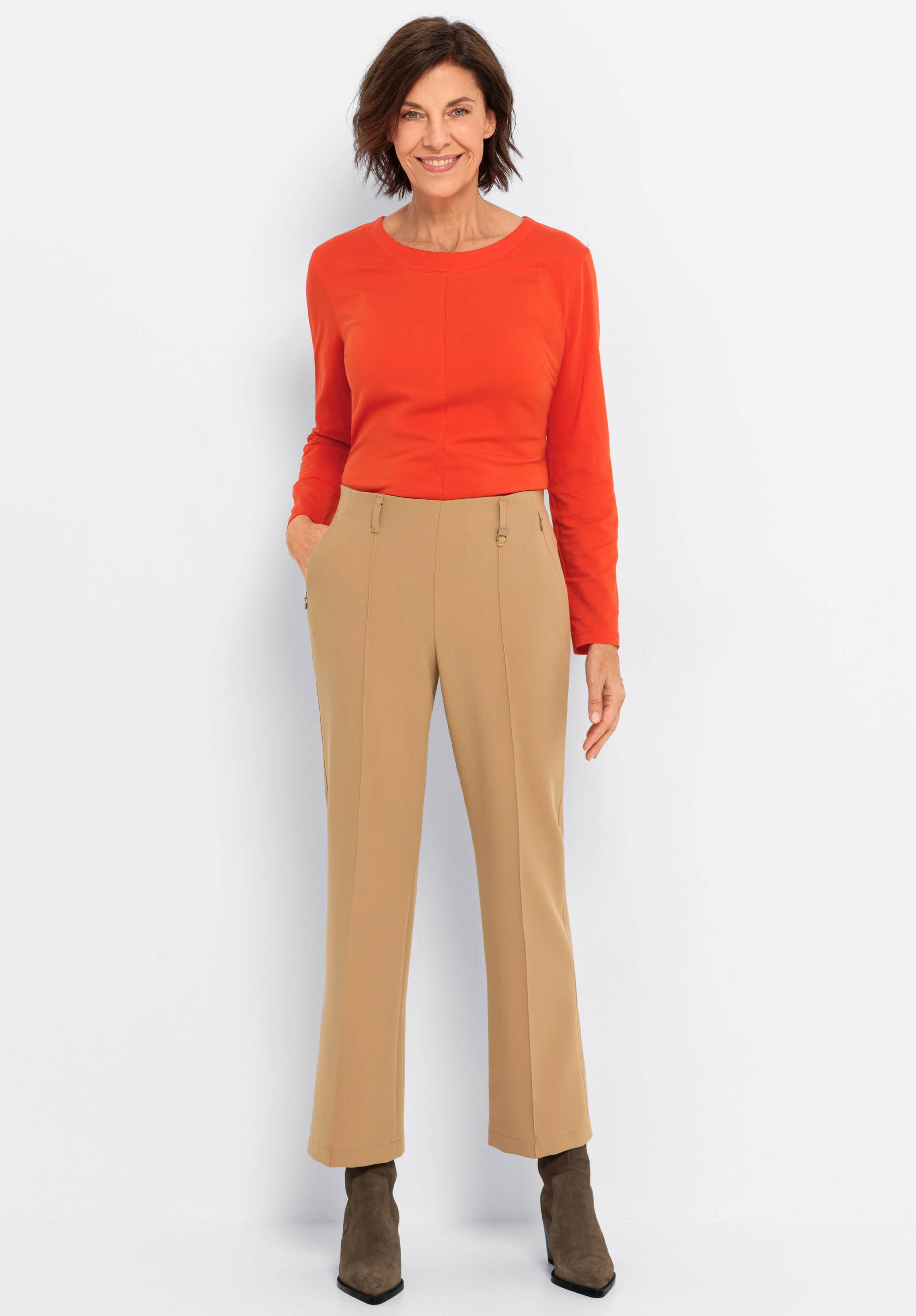 Eine Frau lächelt, während sie vor einem weißen Hintergrund steht. Sie trägt ein langärmeliges orangefarbenes Oberteil, eine hellbraune, hochtaillierte Hose und braune Stiefeletten, die Hände in den Taschen.