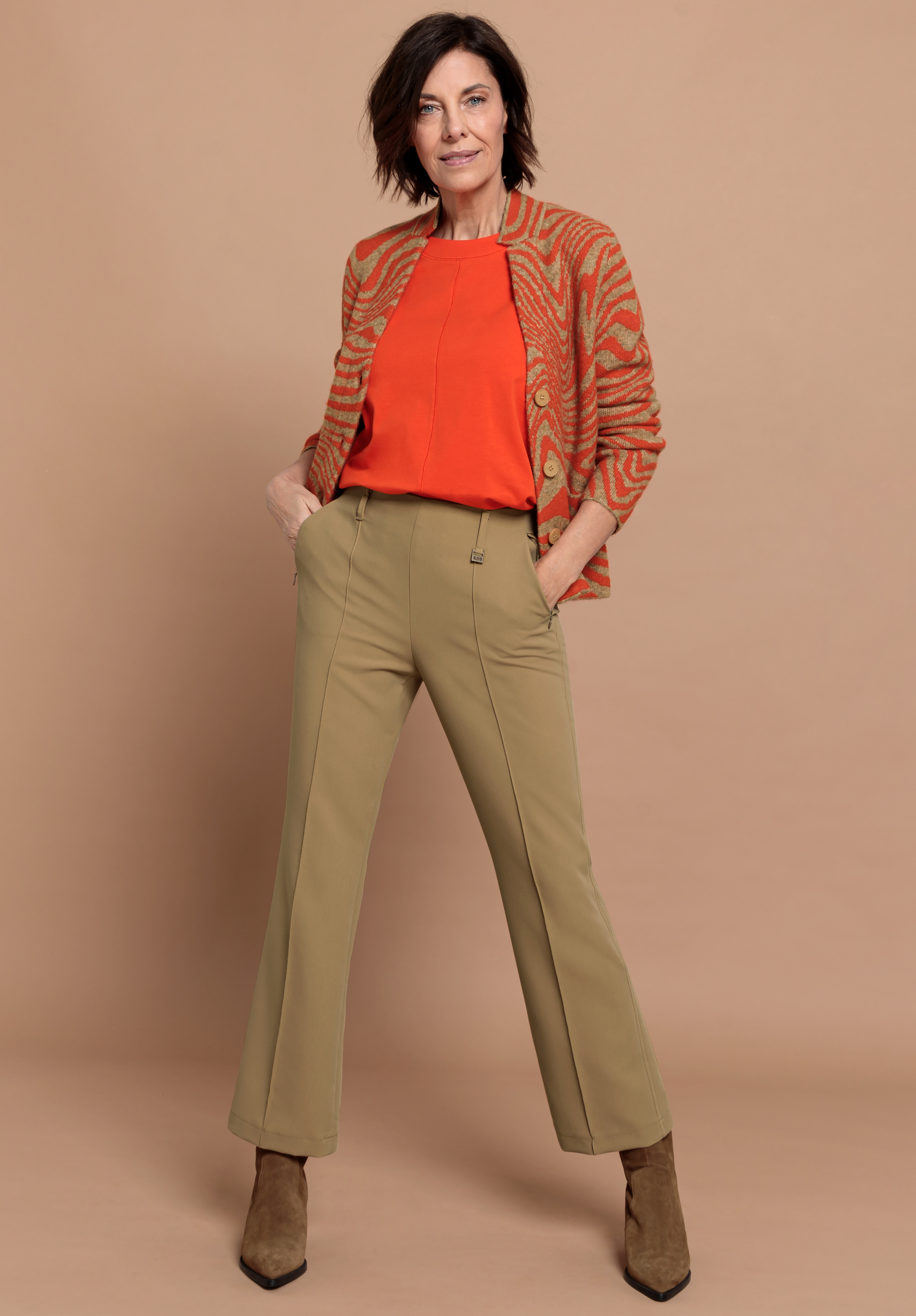 Eine Frau steht selbstbewusst mit den Händen in den Taschen da. Sie trägt ein orangefarbenes Oberteil, eine gemusterte orangefarbene Strickjacke, eine khakifarbene Hose mit weitem Bein und braune Stiefeletten vor einem beigen Hintergrund.