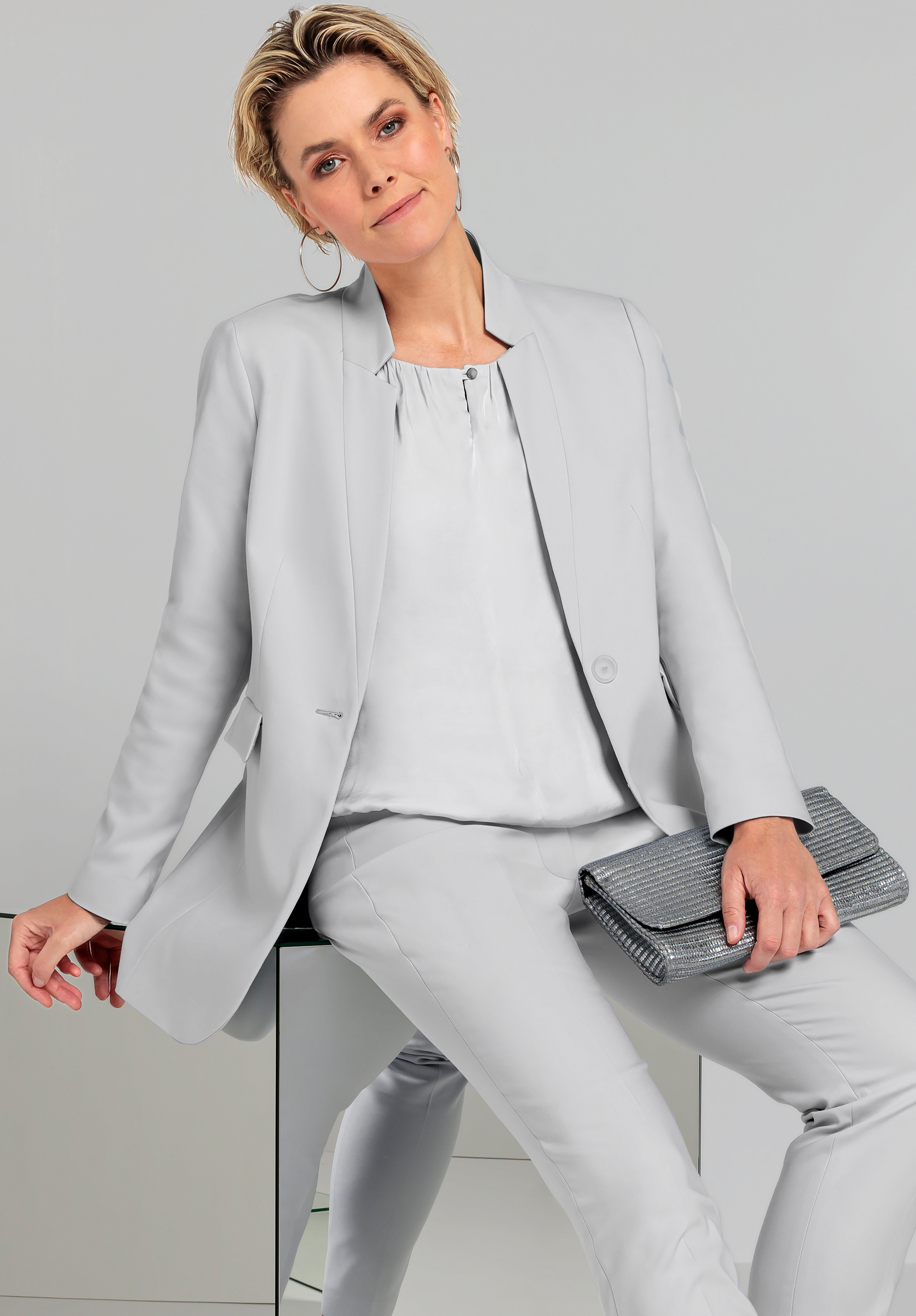 Eine Frau mit kurzen blonden Haaren trägt ein hellgraues Kostüm und hält eine strukturierte Clutch in der Hand. Sie sitzt auf einer reflektierenden Oberfläche und blickt vor einem schlichten grauen Hintergrund selbstbewusst in die Kamera.