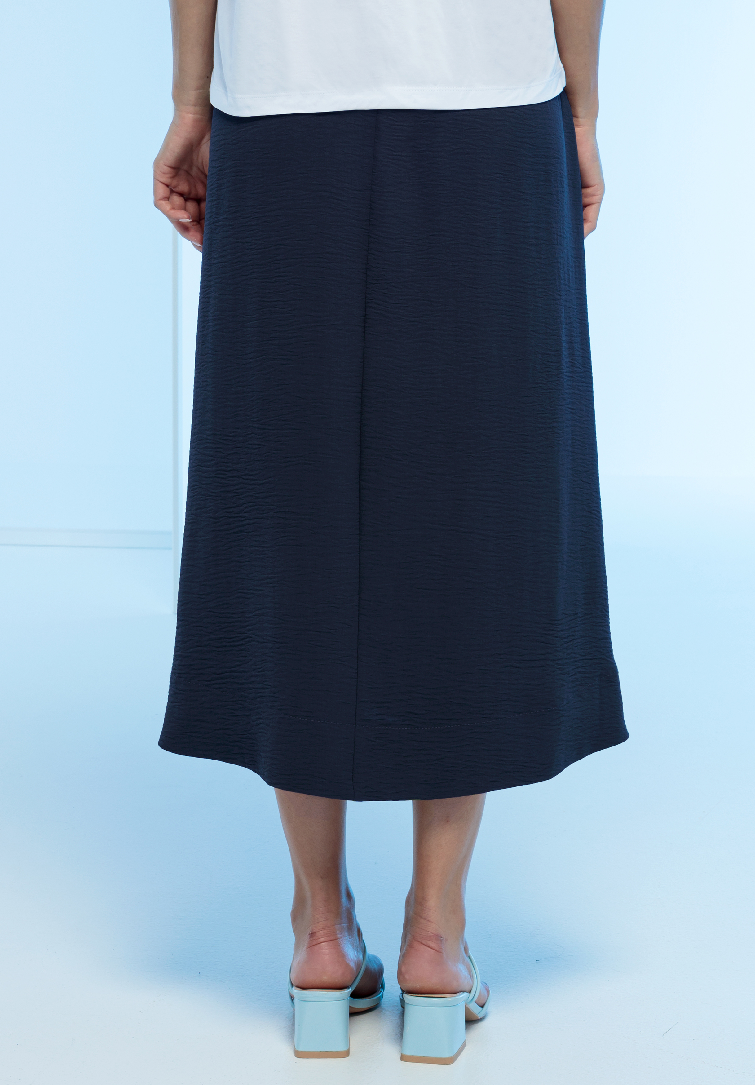 Eine Person steht mit dem Rücken zur Kamera, trägt ein weißes Oberteil, einen marineblauen Midirock und hellblaue Sandalen mit Absatz auf einem hellblauen Boden.