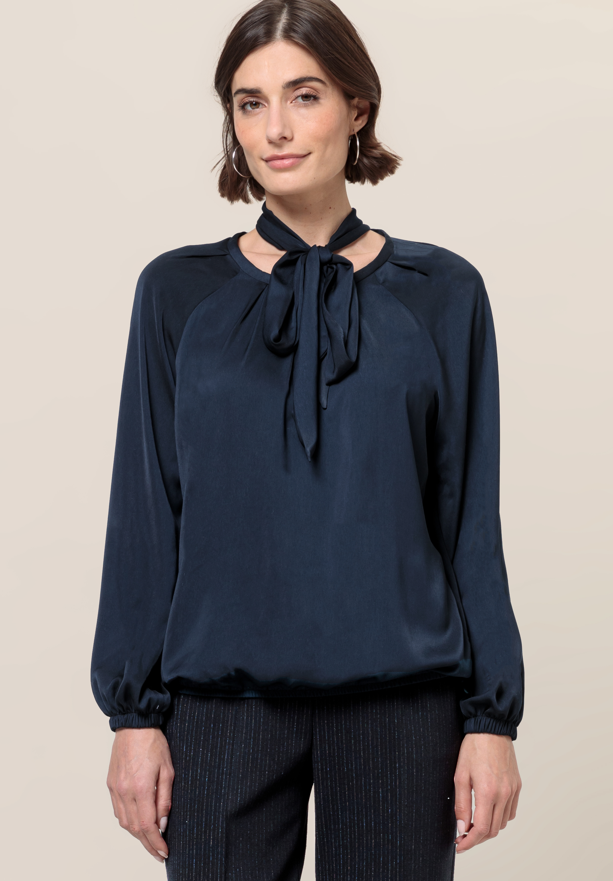 Eine Frau mit kurzen braunen Haaren trägt eine dunkelblaue Langarmbluse mit Schleife am Kragen und eine schwarze Hose. Sie steht vor einem schlichten hellbeigen Hintergrund und blickt mit neutralem Gesichtsausdruck geradeaus.