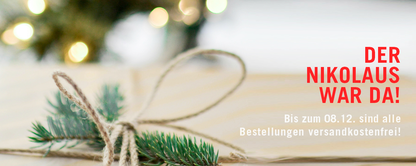 Ein verpacktes Geschenk mit Tannenzweigen und Bindfäden vor verschwommener Weihnachtsbeleuchtung. Der Text auf Deutsch lautet: „Der Nikolaus war da! Bis zum 08.12. sind alle Bestellungen versandkostenfrei!“.