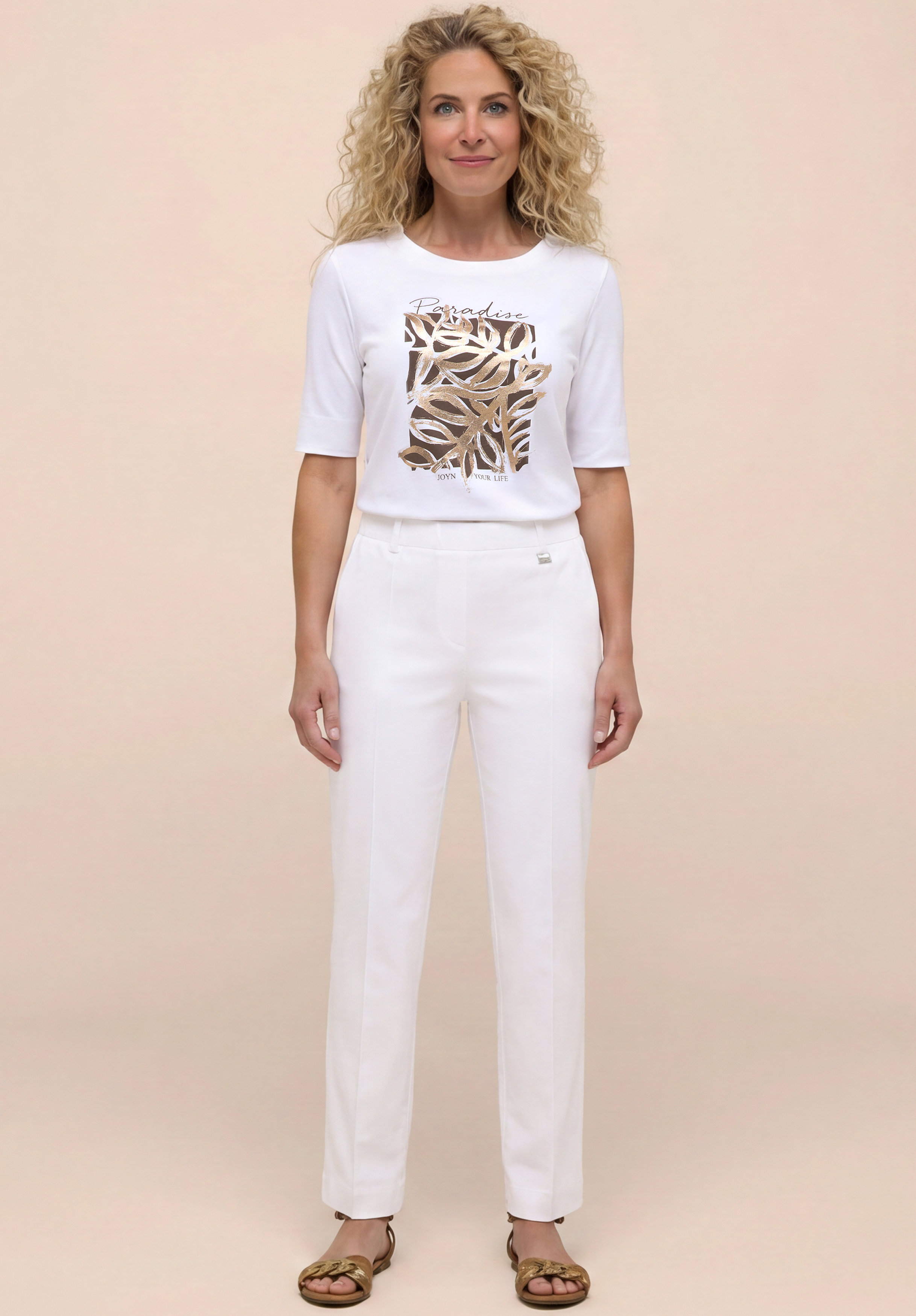 Eine Frau mit lockigem blondem Haar steht da und lächelt. Sie trägt ein weißes T-Shirt mit einem abstrakten Muster in Gold und Braun, eine weiße Hose und goldene Sandalen vor einem hellbeigen Hintergrund.