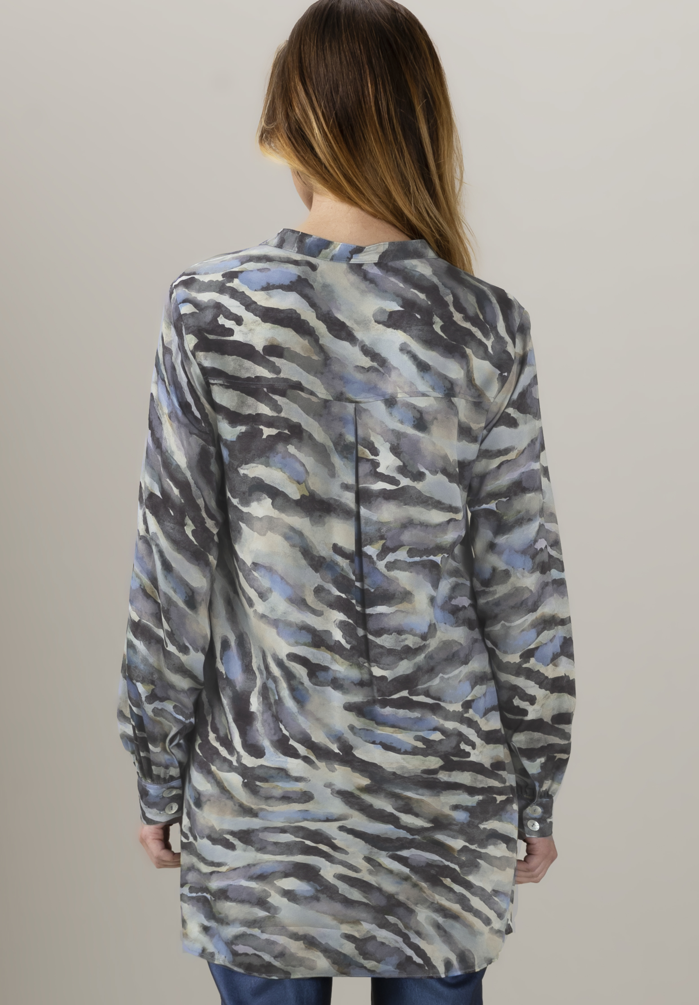 Eine Frau mit langen braunen Haaren, von hinten gesehen, trägt eine weite, langärmelige Tunika mit einem abstrakten Muster in Blau, Grau und Schwarz über Blue Jeans vor einem schlichten hellen Hintergrund.