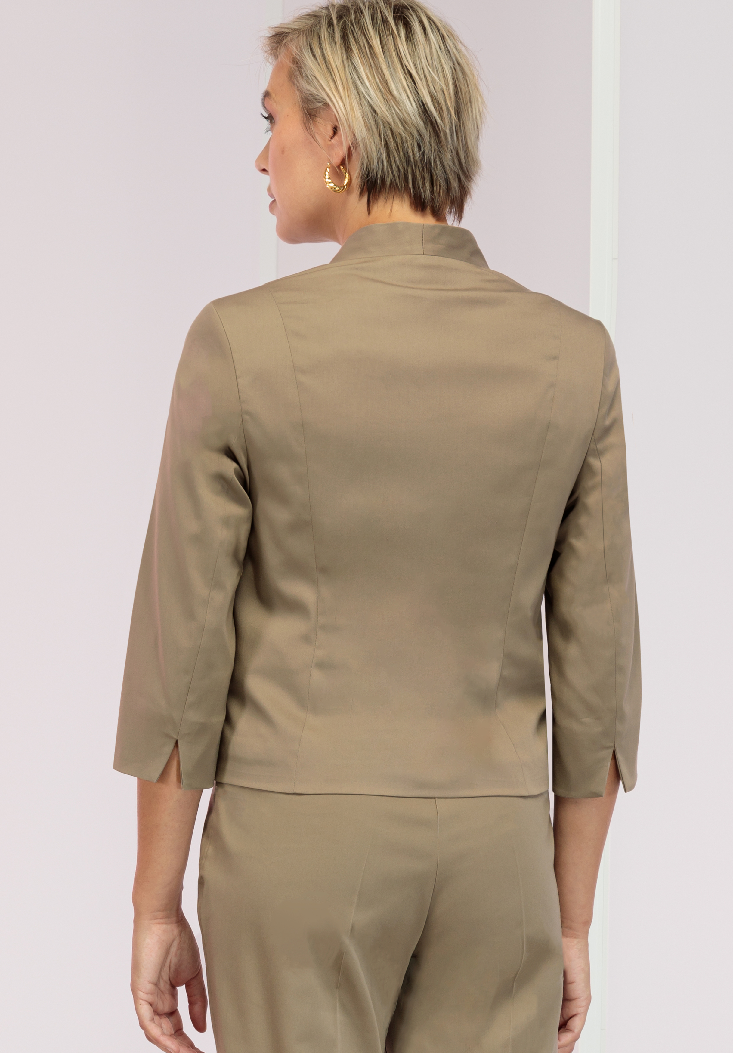 Eine Person mit kurzen blonden Haaren, goldenen Creolen und einem hellbraunen Maßanzug ist von hinten zu sehen. Das Jackett hat Dreiviertelärmel mit kleinen Schlitzen.