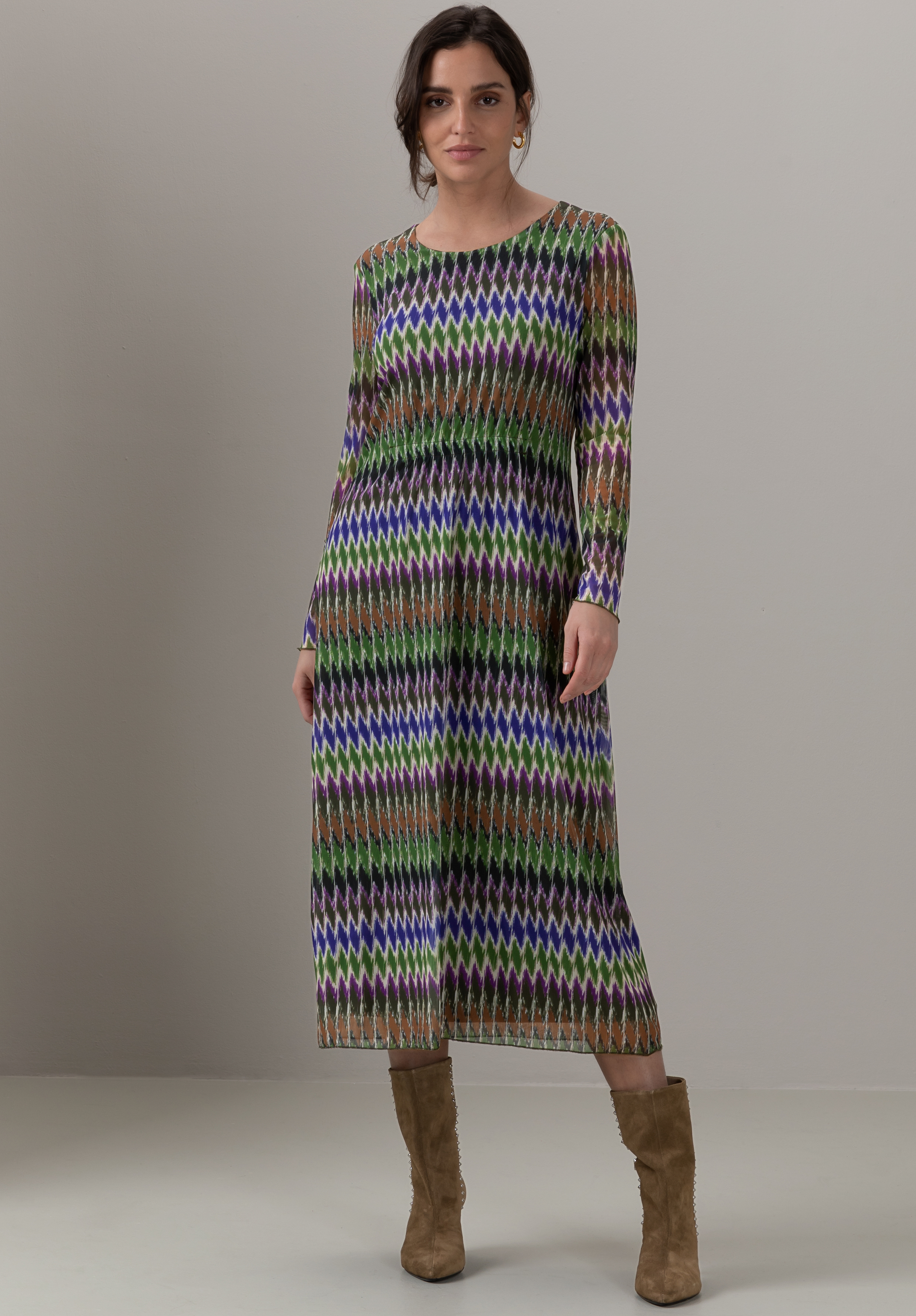 Eine Frau in einem langärmeligen, knöchellangen Kleid mit buntem Zickzackmuster in Grün, Lila und Braun steht vor einem schlichten Hintergrund. Sie hat kurzes dunkles Haar und trägt hellbraune Wildlederstiefel.
