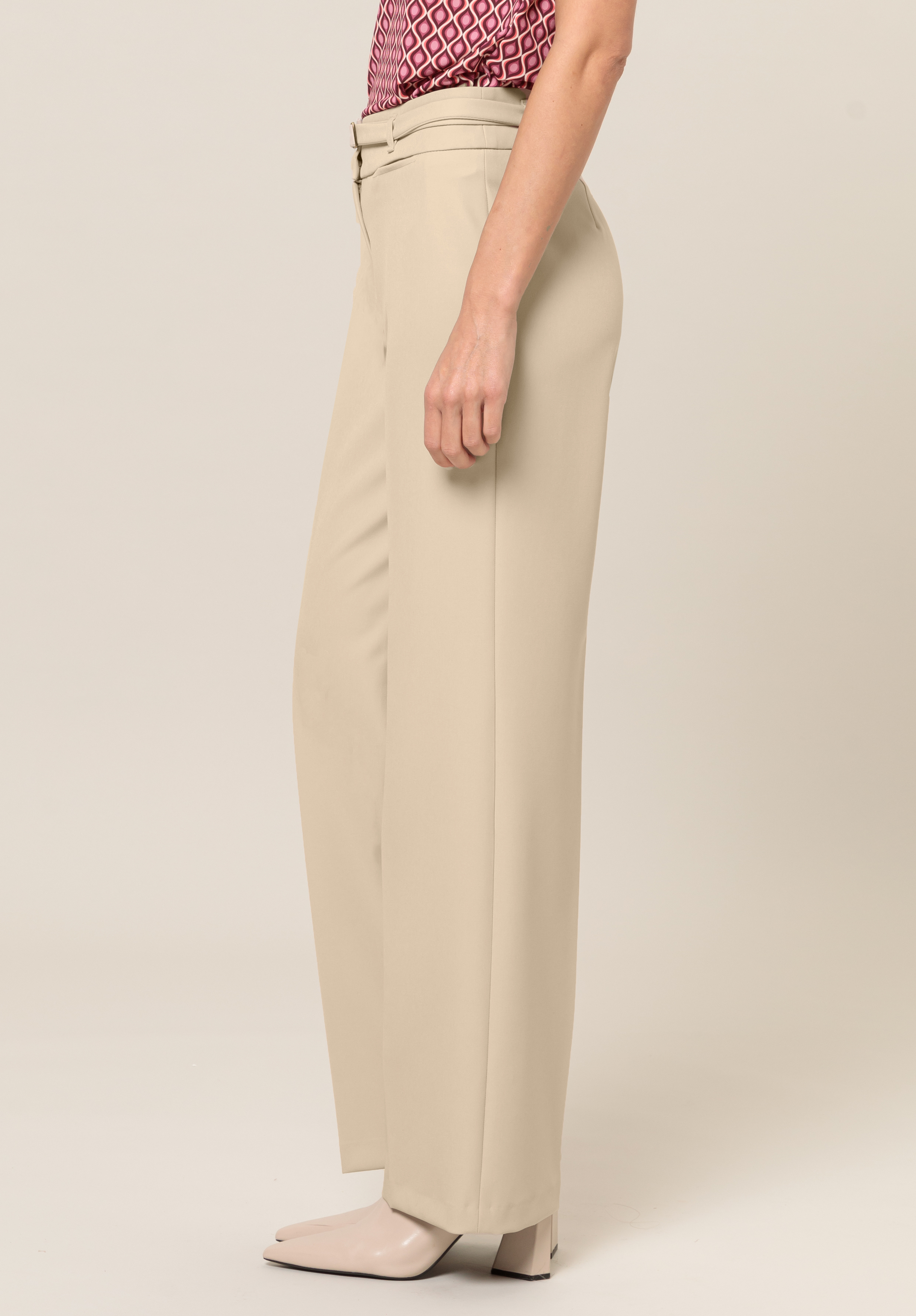 Eine Person trägt hoch taillierte, beigefarbene Hosen mit weitem Bein, ein gemustertes rosa Oberteil und helle spitze Schuhe und steht im Profil vor einem schlichten Hintergrund.
