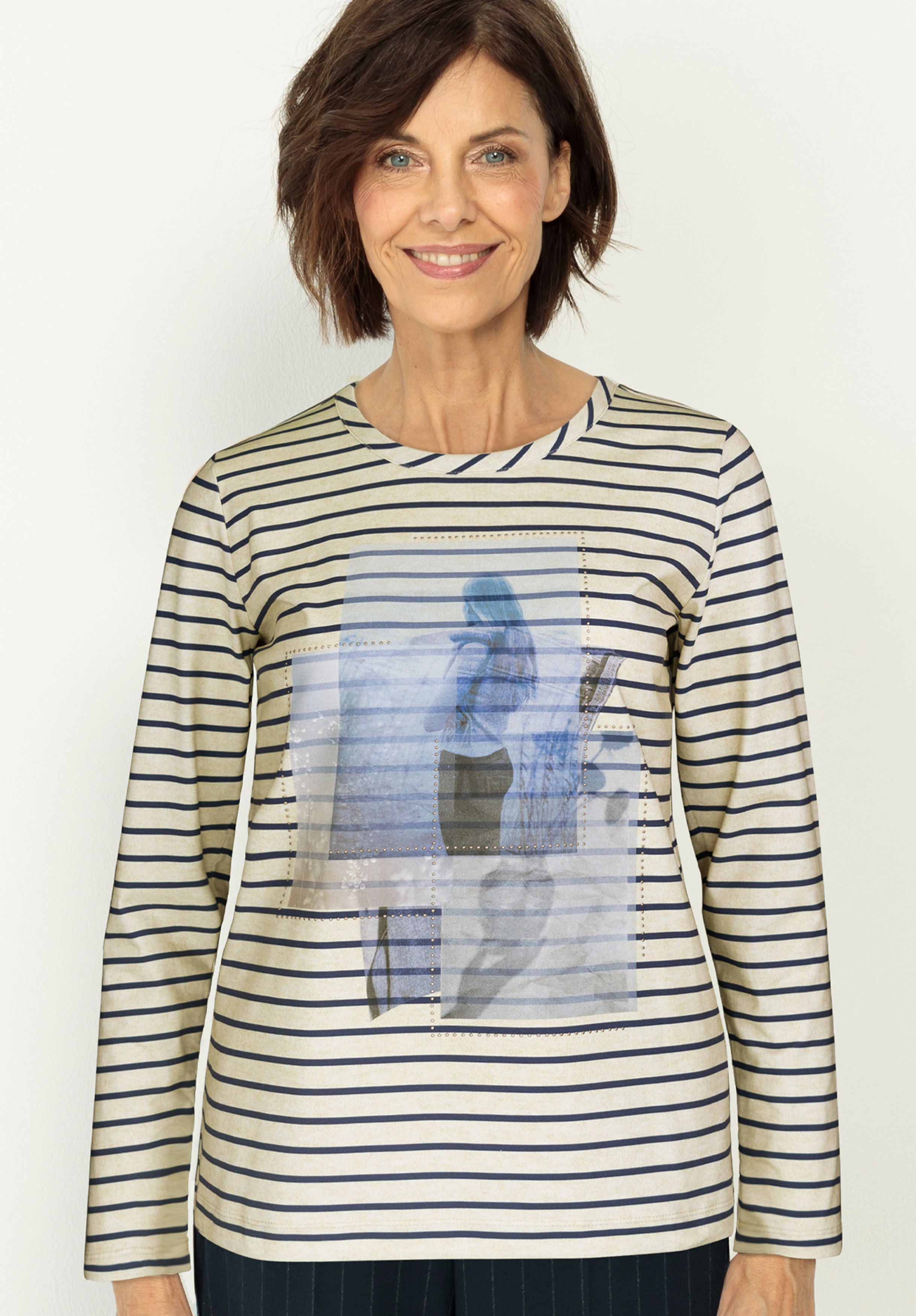 Eine Frau mit kurzen braunen Haaren lächelt, sie trägt ein langärmeliges, cremefarben und marineblau gestreiftes Hemd mit rechteckigen Aufnäher mit einem abstrakten, blau getönten Bild auf der Vorderseite. 