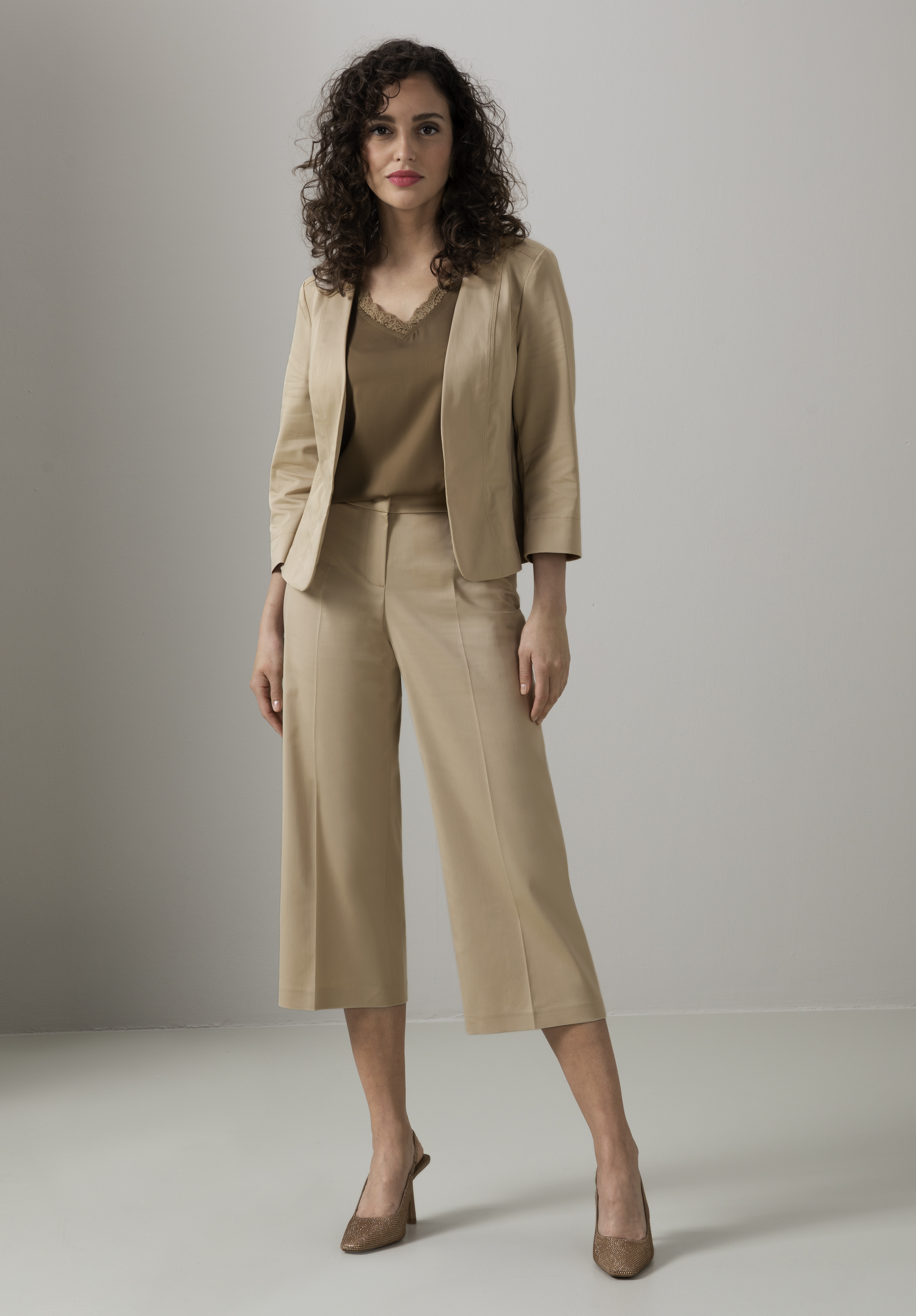 Eine Frau mit lockigem Haar steht vor einem schlichten Hintergrund. Sie trägt einen hellbraunen Blazer, kurze hellbraune Hosen, ein braunes Oberteil und braune Pumps. Sie blickt mit neutralem Gesichtsausdruck nach vorne.