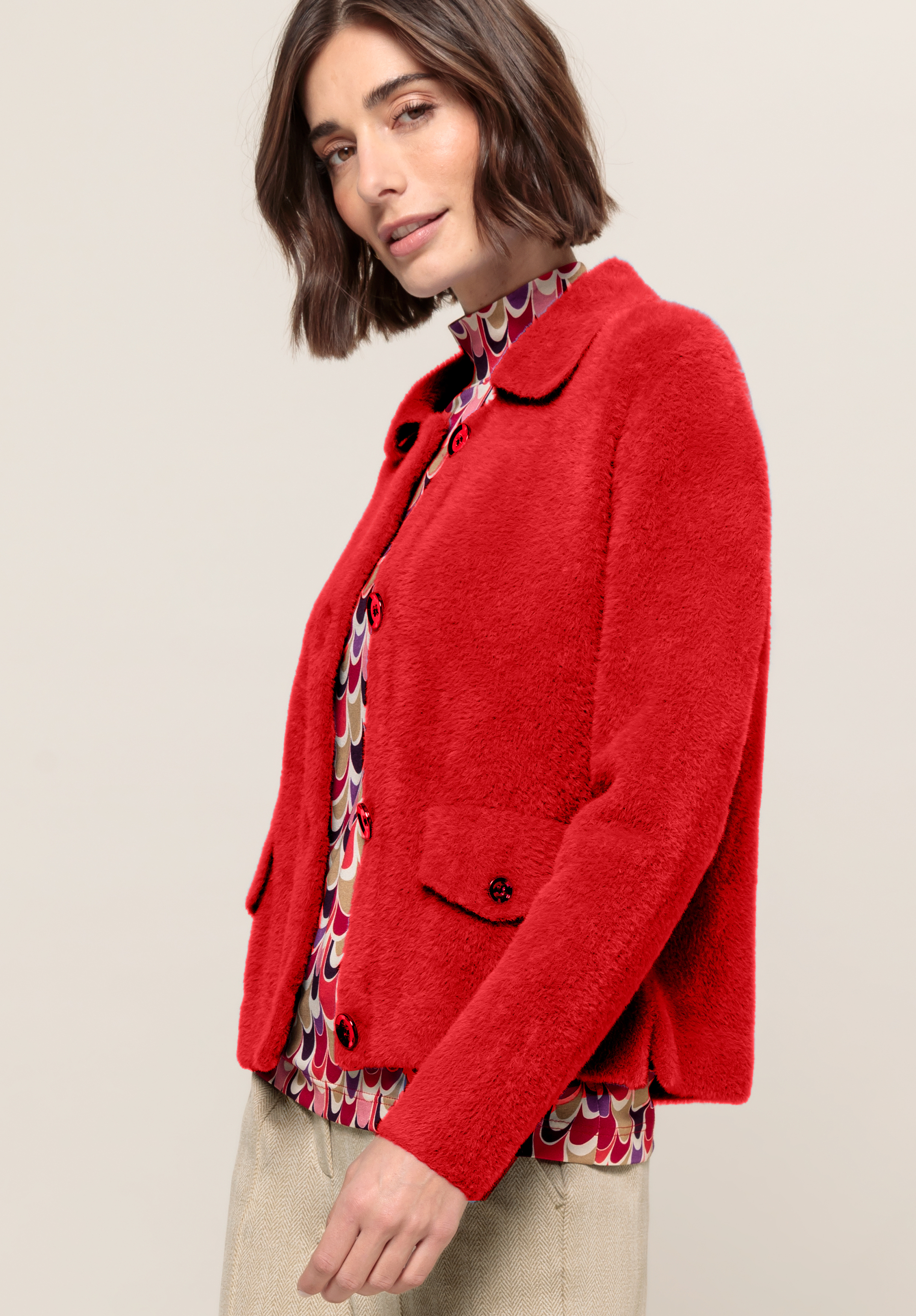 Frau mit Bob-Haarschnitt trägt eine leuchtend rote Strukturjacke über einer bunt gemusterten Bluse und beigen Hosen und steht vor einem neutralen Hintergrund. Sie ist leicht zur Seite gewandt und blickt in die Kamera.