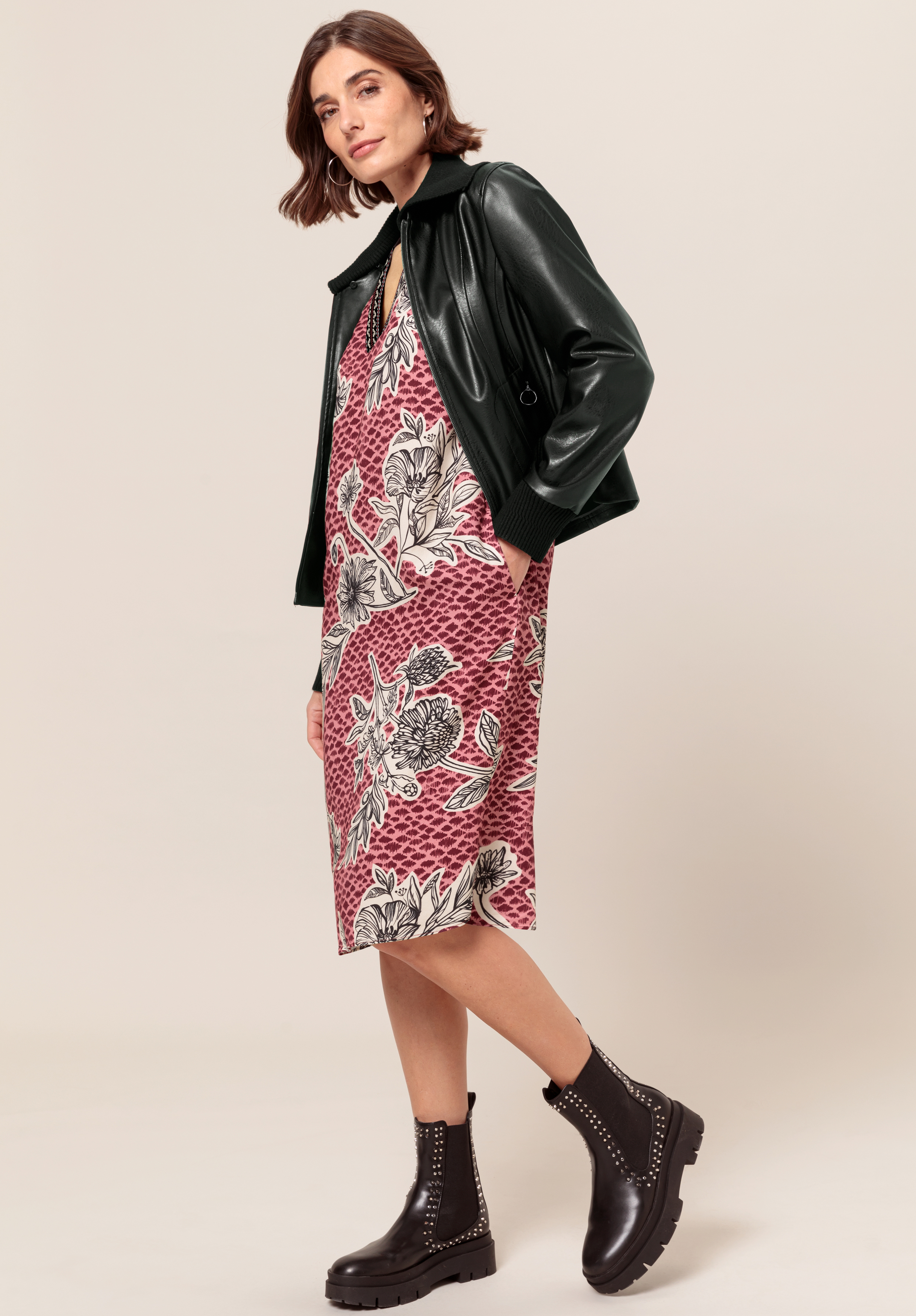 Eine Frau posiert in einem rosa-weißen Blumenkleid mit einer schwarzen Lederjacke und klobigen schwarzen Stiefeln mit Nieten vor einem schlichten beigen Hintergrund.