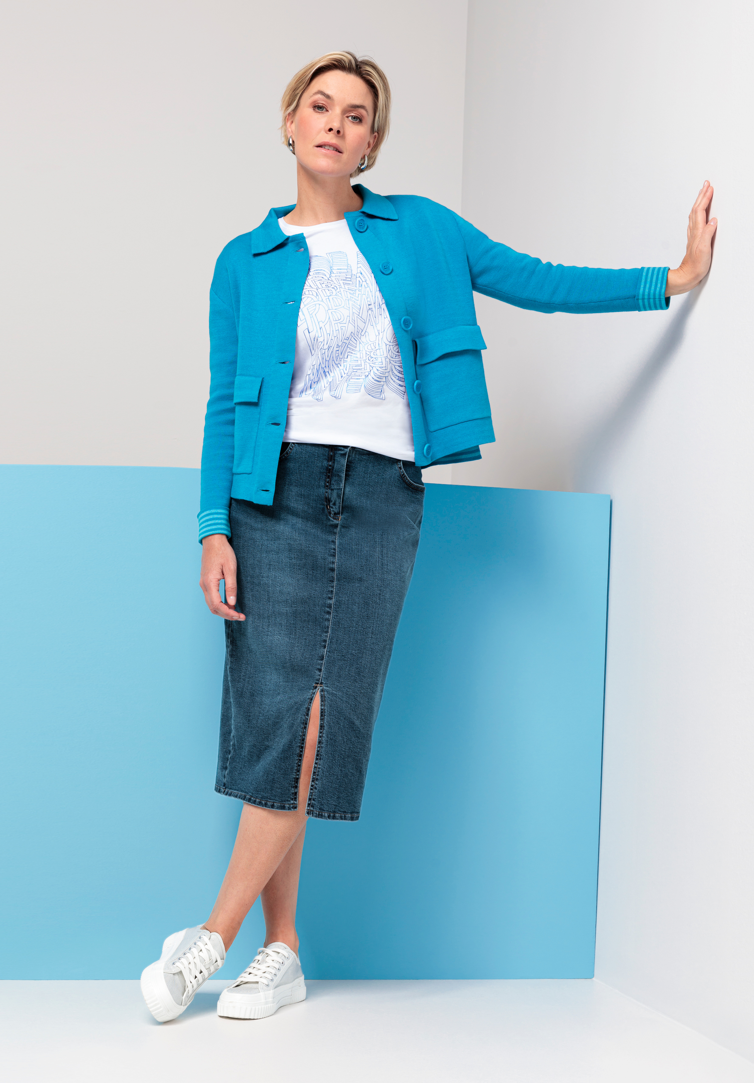 Vor einem weiß-blauen Hintergrund steht eine Frau, sie trägt eine leuchtend blaue Jacke, ein weißes bedrucktes T-Shirt, einen knielangen Jeansrock mit Schlitz vorne und weiße Plateau-Sneakers.