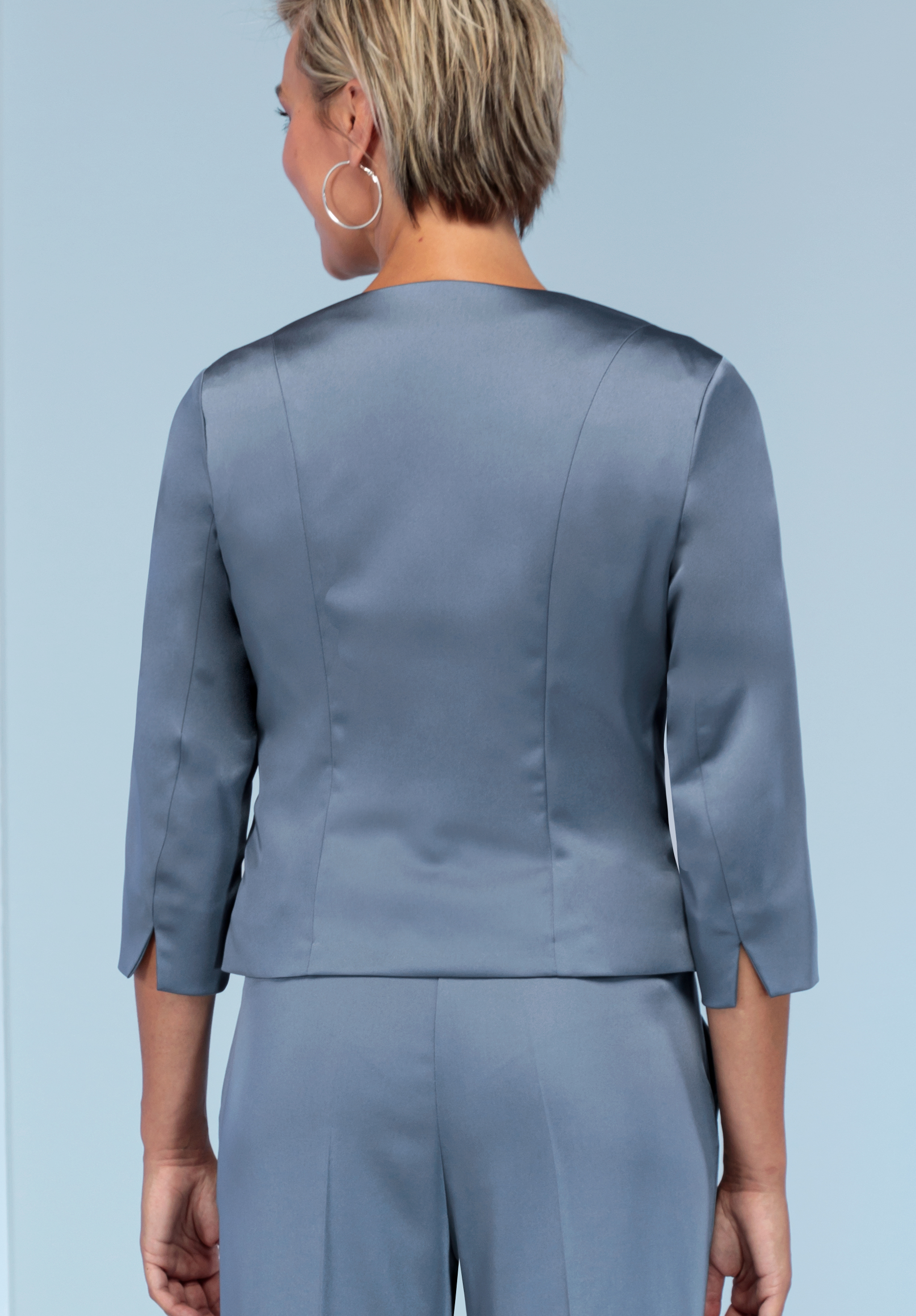 Eine Frau mit kurzen blonden Haaren, großen Creolen und einem glänzenden grauen Anzug mit Dreiviertelärmeln steht mit dem Rücken zur Kamera vor einem hellblauen Hintergrund.