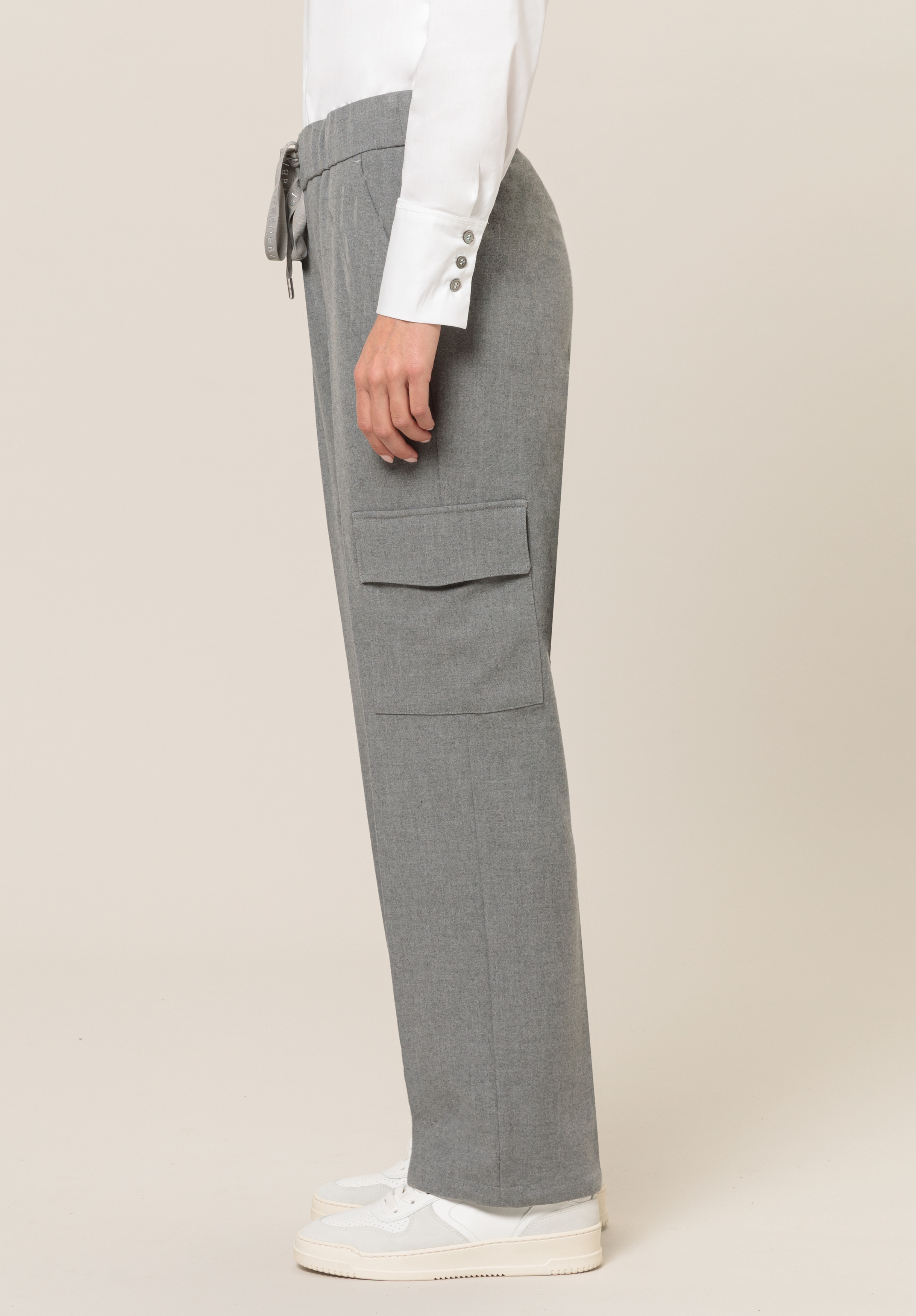 Eine Person trägt hellgraue Cargohosen mit einer großen Seitentasche, ein weißes Hemd mit geknöpften Manschetten und weiße Turnschuhe und steht im Profil vor einem schlichten hellen Hintergrund.