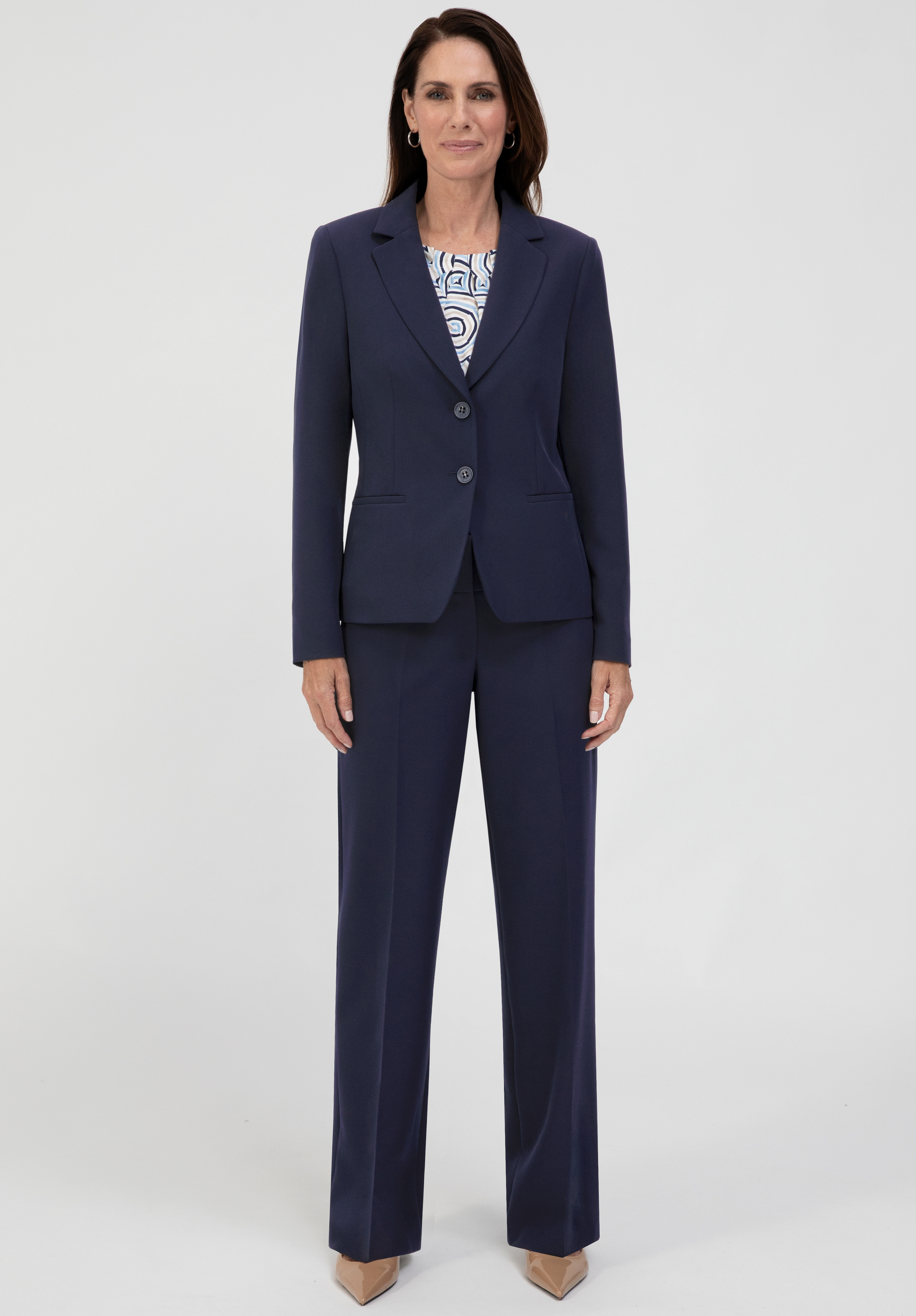 Eine Frau steht mit dem Blick nach vorne und trägt einen marineblauen Business-Anzug, darunter ein gemustertes weiß-blaues Oberteil. Sie hat schulterlanges dunkles Haar und trägt beige Schuhe mit spitzer Zehenpartie. Der Hintergrund ist einfarbig weiß.