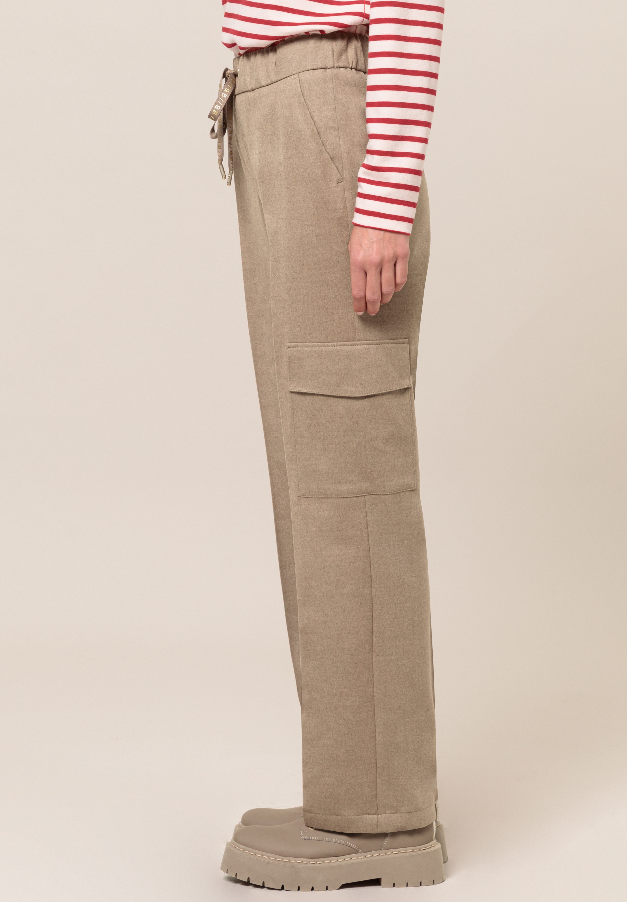 Im Profil steht eine Person, die taupefarbene Cargohosen, ein rot-weiß gestreiftes Langarmhemd und klobige beige Schuhe vor einem schlichten Hintergrund trägt.