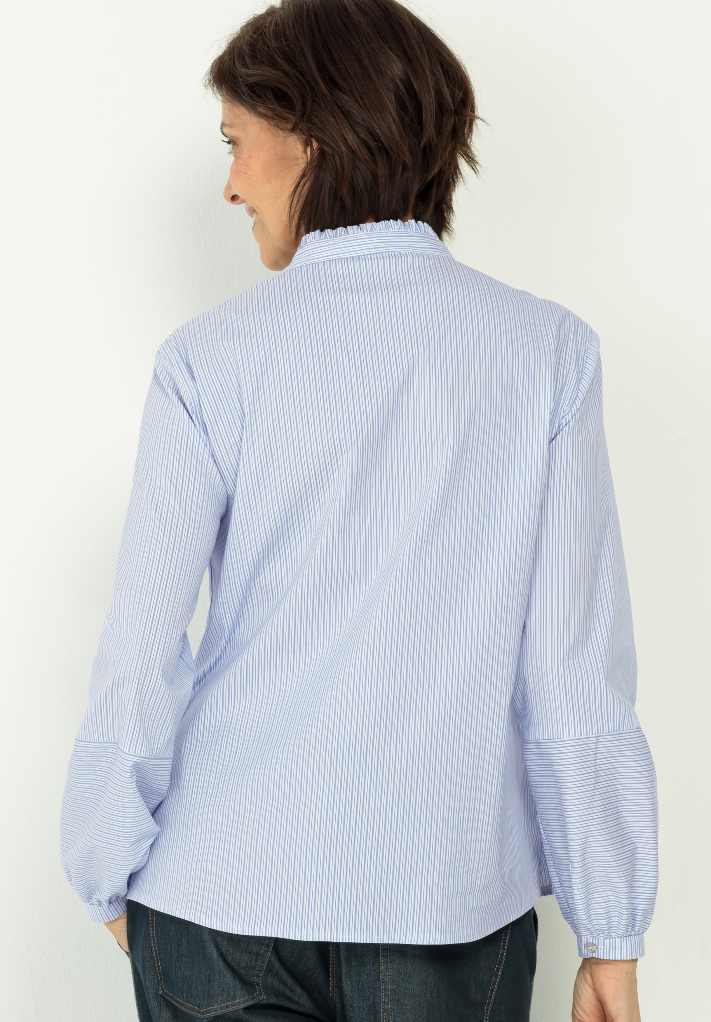 Eine Frau mit kurzen braunen Haaren, von hinten gesehen, trägt eine hellblau-weiß gestreifte langärmelige Bluse mit Rüschenkragen und eine dunkle Hose vor einem hellen Hintergrund.