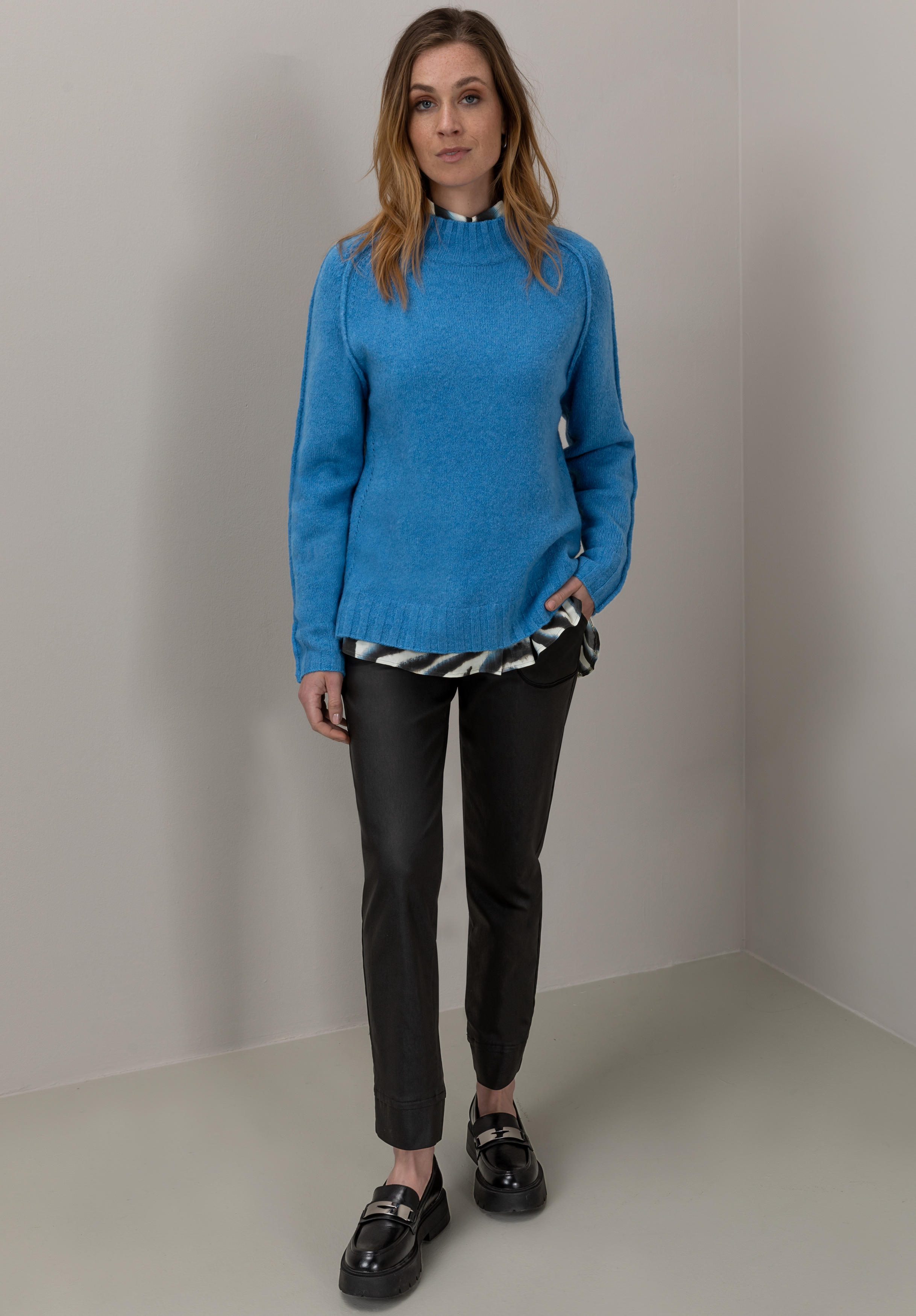 Eine Frau steht in einer Ecke. Sie trägt einen blauen Pullover über einem gestreiften Hemd, eine schwarze Hose und schwarze Slipper. Sie hat schulterlanges Haar, einen neutralen Gesichtsausdruck und eine Hand in der Tasche.