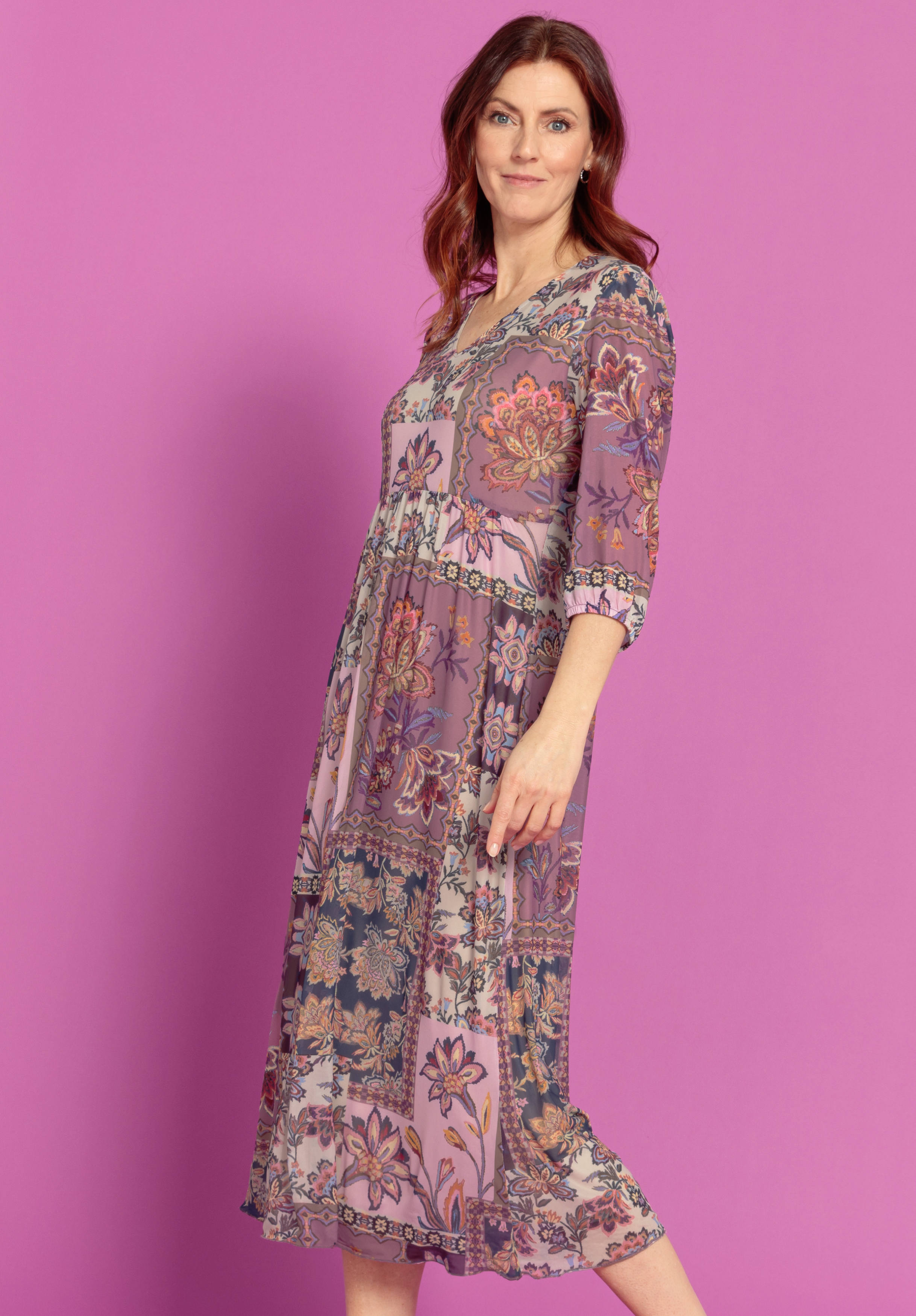 Eine Frau mit gewelltem rotem Haar trägt ein langes, blumengemustertes Kleid in Lila- und Rosatönen. Sie steht vor einem einfarbigen rosa Hintergrund und lächelt leicht, während sie im Profil posiert.